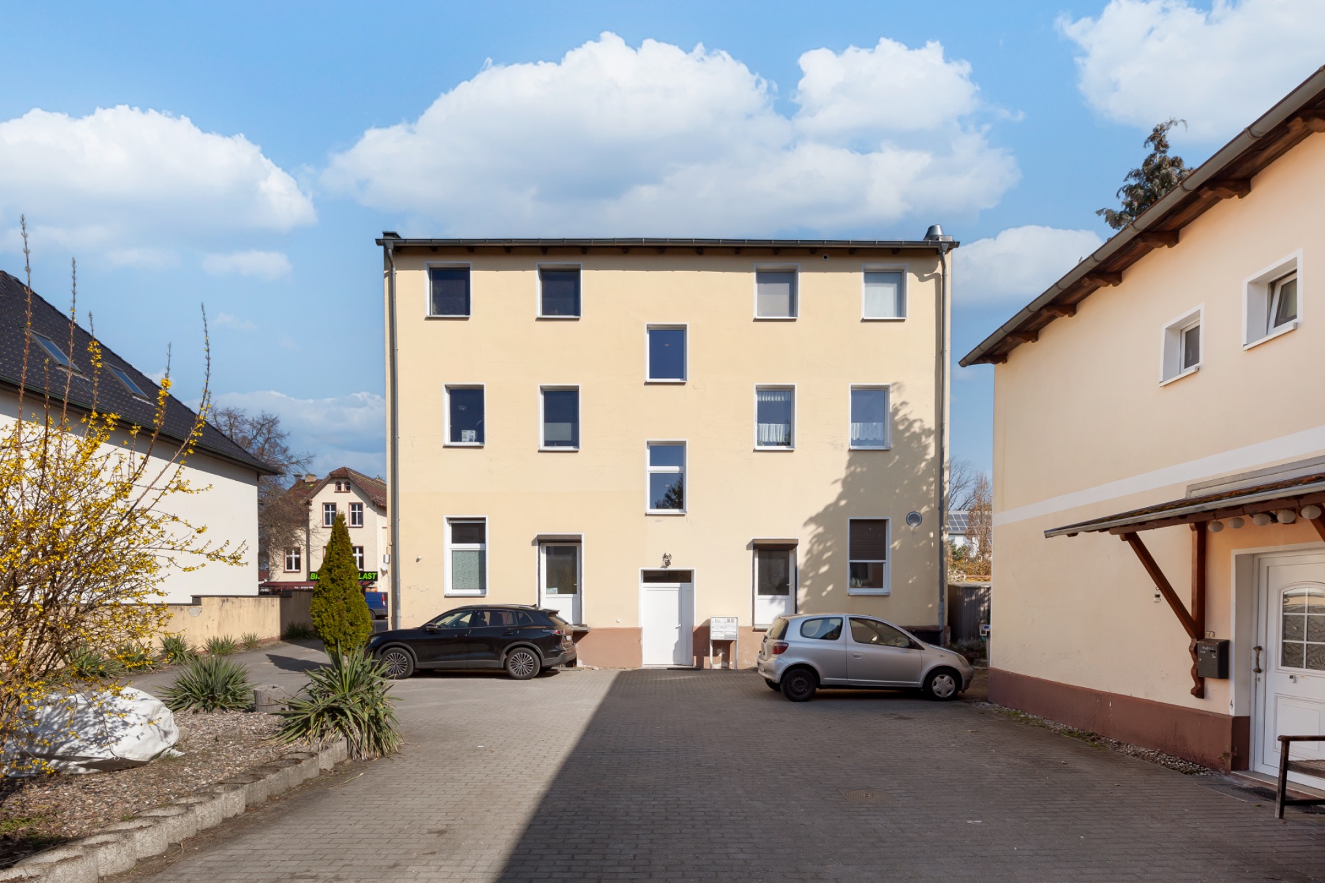 Dreistöckiges Wohnhaus mit sechs Fenstern und zwei geparkten Autos im Hof.