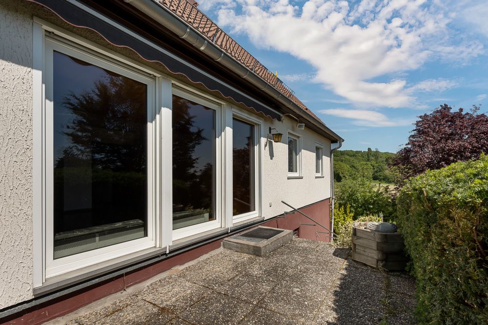 Terrasse mit großen Fenstern an einem Wohnhaus, umgeben von Büschen und Bäumen bei sonnigem Wetter.
