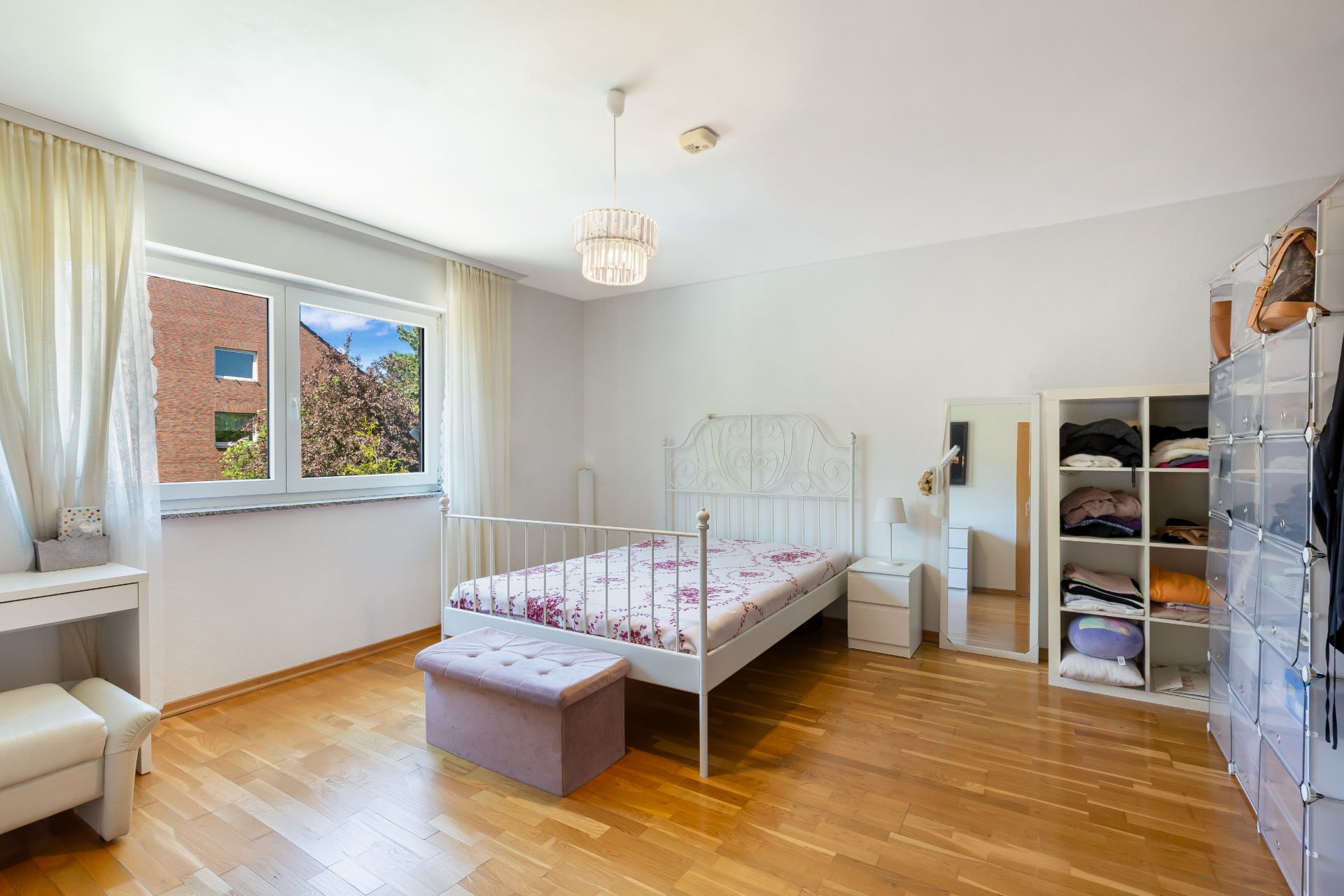 Helles Schlafzimmer mit Bett, Kleiderschrank, Spiegel und Schreibtisch vor einem Fenster.