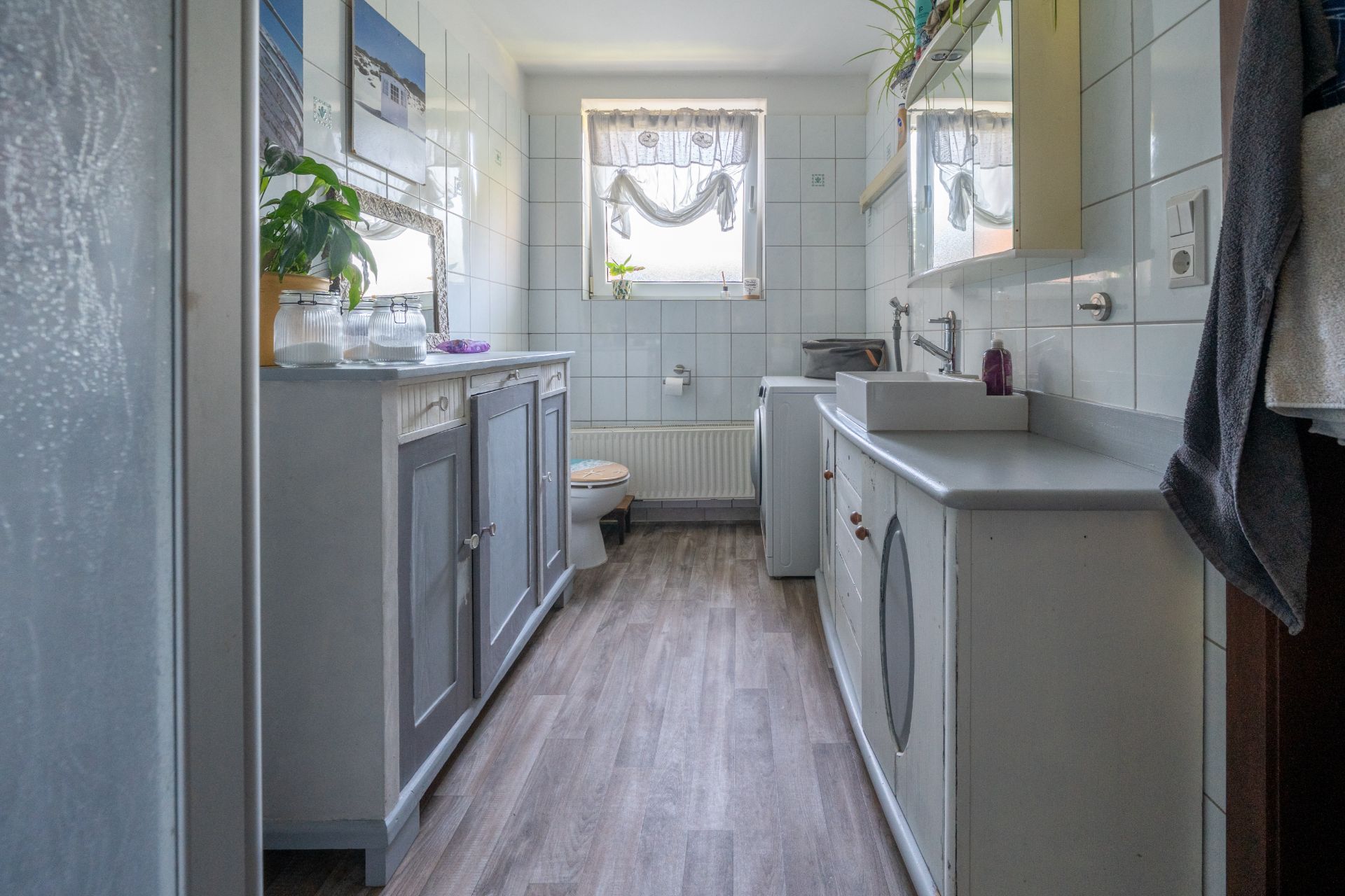 Helles Badezimmer mit Fenster, Waschbecken, Schränken, Spiegel und Holzboden.