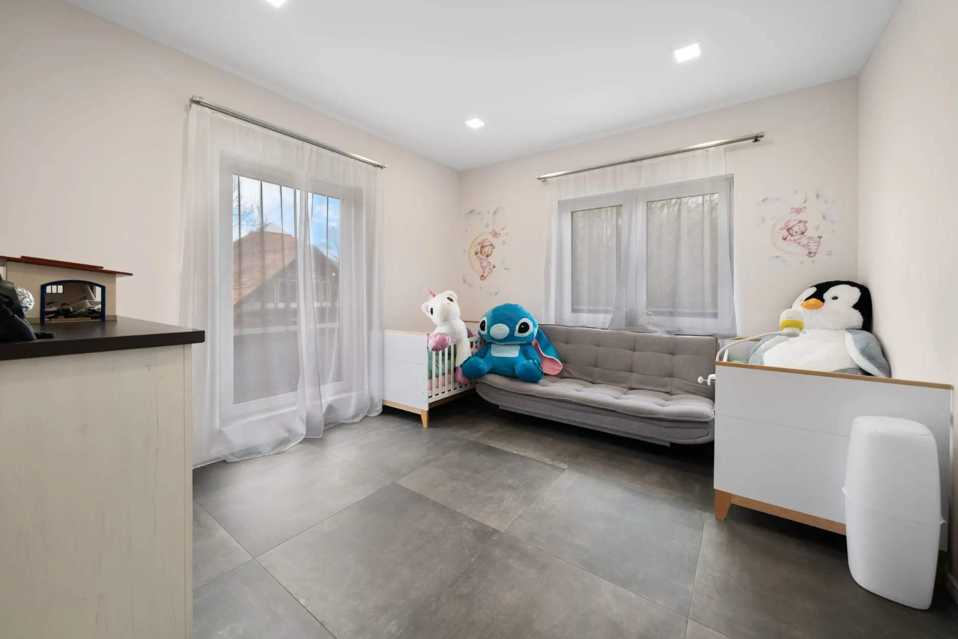 Helles Kinderzimmer mit Sofa, Babybett, Kommode und großen Kuscheltieren.