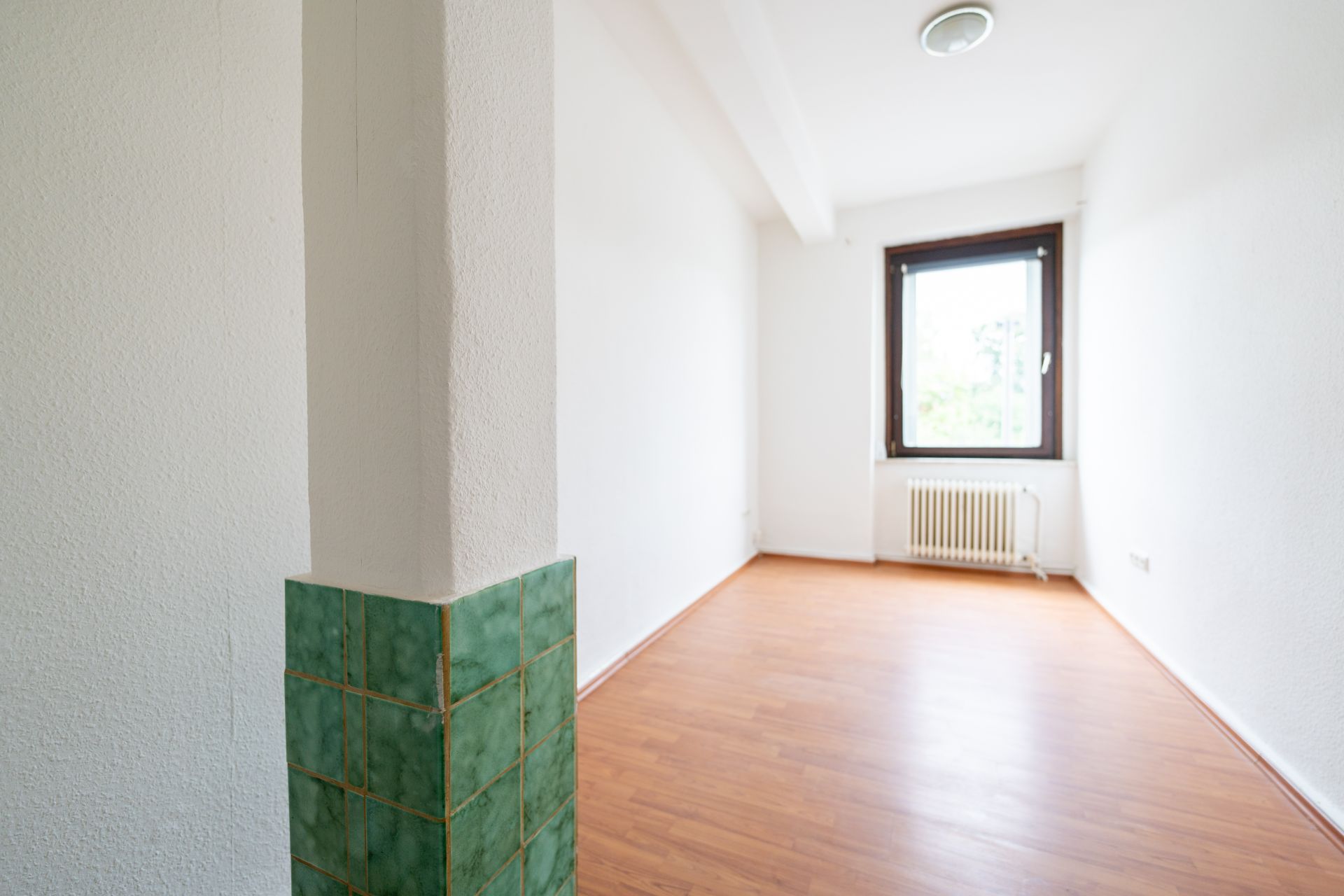 Leerer Raum mit weißer Wand, Holzfußboden, Fenster und Heizkörper, grün gefliester Pfeiler im Vordergrund.