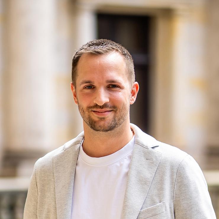 Ein Mann mit kurzem Haar und Bart trägt ein helles Sakko und ein weißes T-Shirt, steht im Freien.