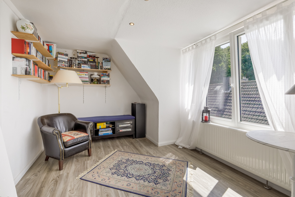 Helles Zimmer mit Sessel, Teppich, Bücherregal, Bank und großen Fenstern mit weißen Vorhängen.