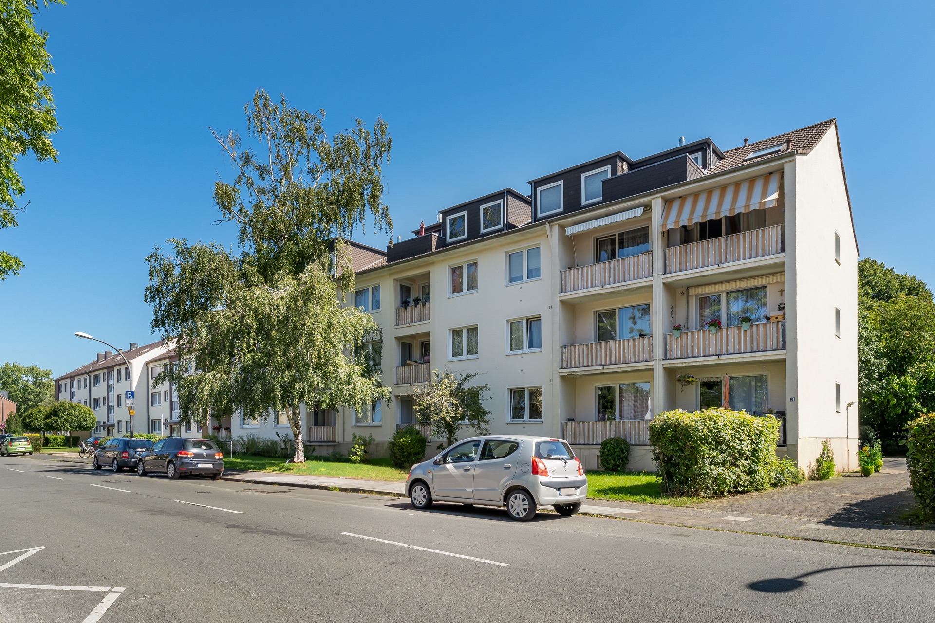 Mehrfamilienhaus mit Balkonen und geparkten Autos an einer ruhigen Straße bei sonnigem Wetter.
