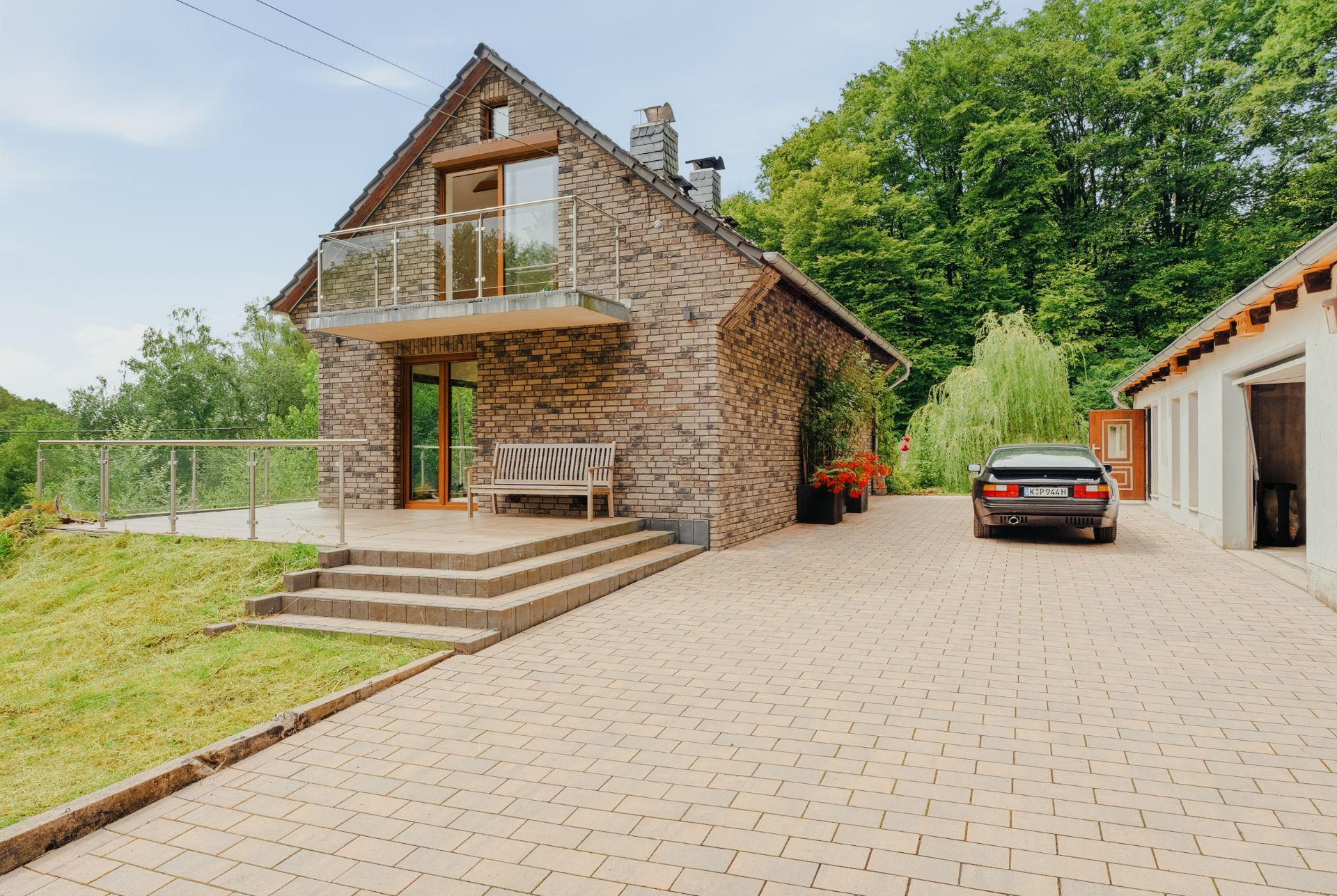 Steinhaus mit Balkon, Terrasse und Sitzbank, daneben gepflasterte Auffahrt mit geparktem Auto.