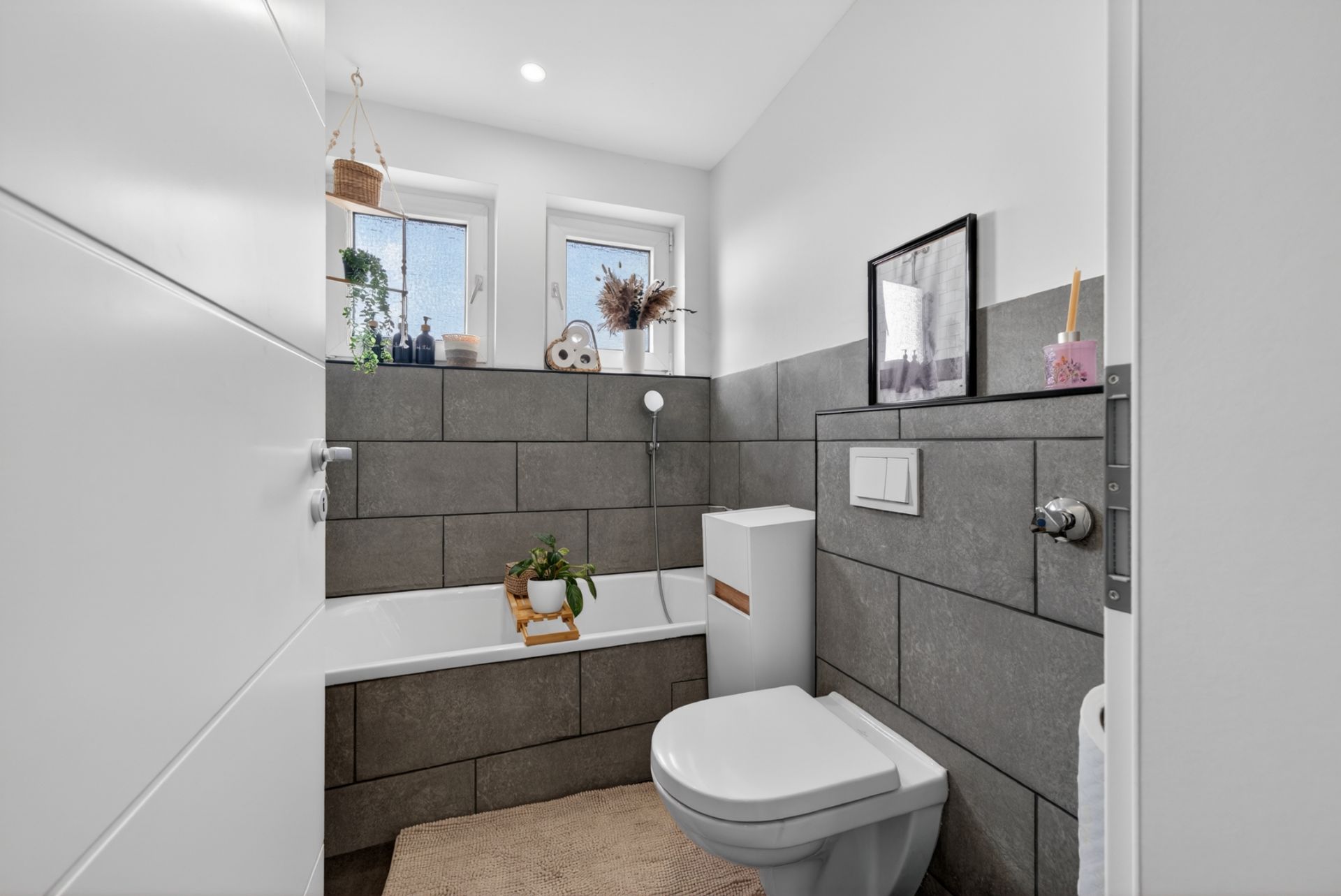 Modernes Badezimmer mit Badewanne, Toilette, grauen Fliesen und zwei Fenstern.