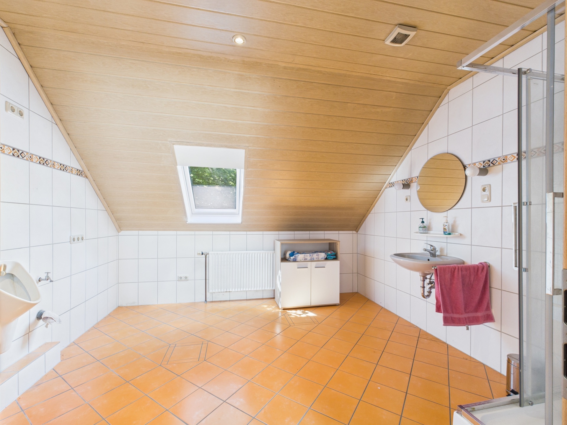 Helles Badezimmer mit schräger Holzdecke, Fenster, Waschbecken, Dusche und Fliesenboden.