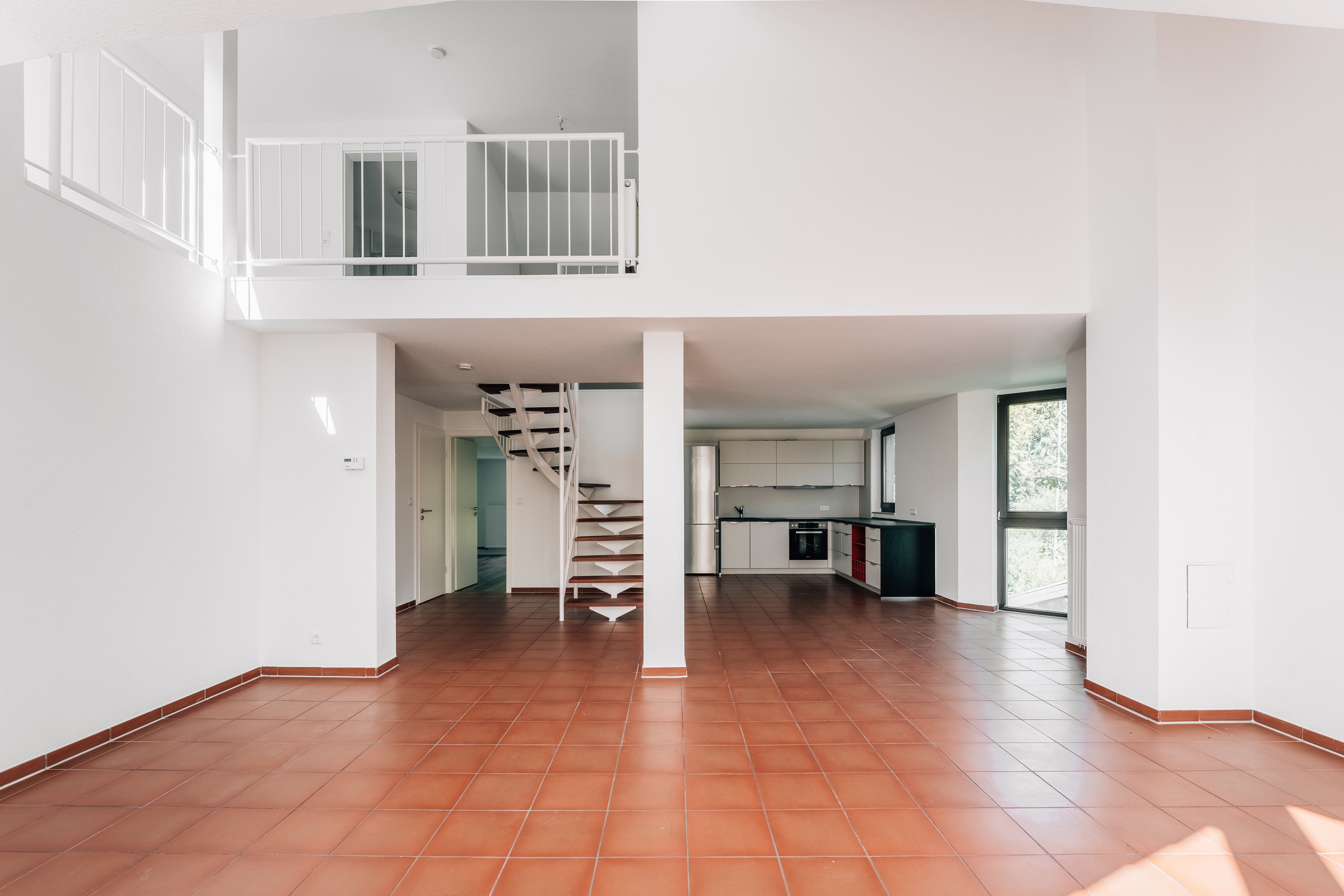 Heller, leerer Wohnraum mit rotem Fliesenboden, offener Küche und Wendeltreppe.