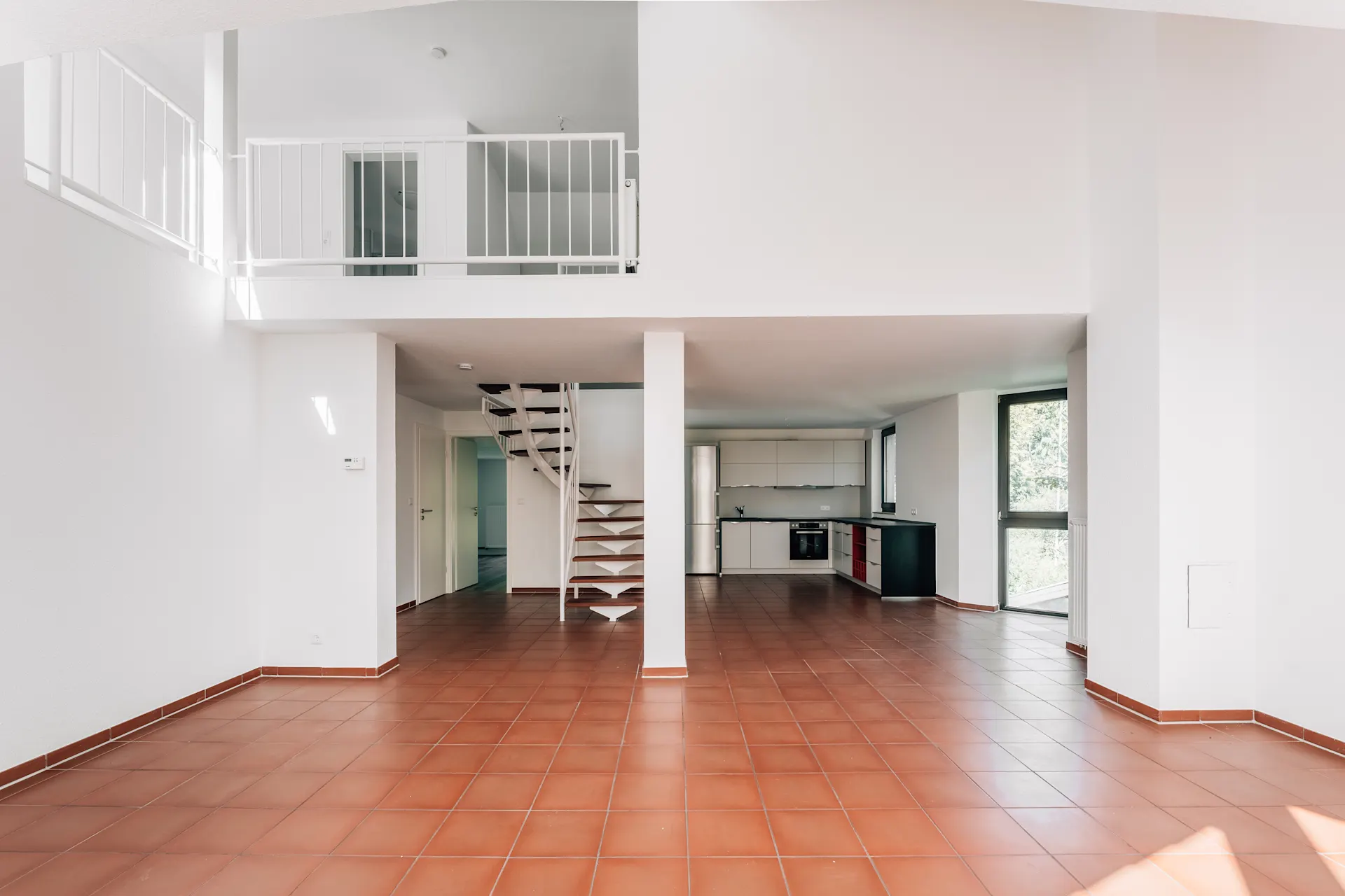 Heller, leerer Wohnraum mit rotem Fliesenboden, offener Küche und Wendeltreppe.