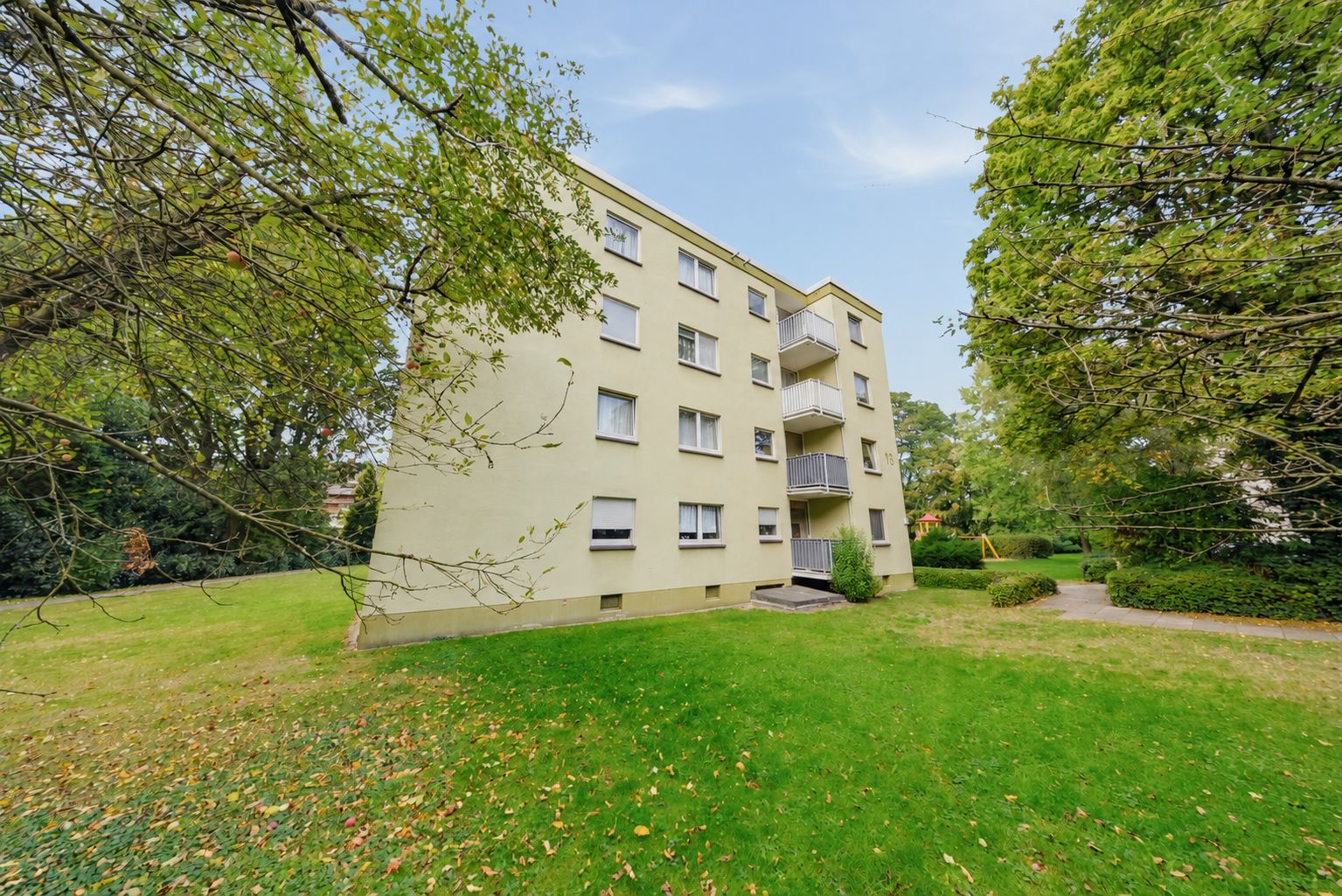 Vierstöckiges Wohnhaus mit Balkonen und gepflegtem grünen Garten, umgeben von Bäumen.