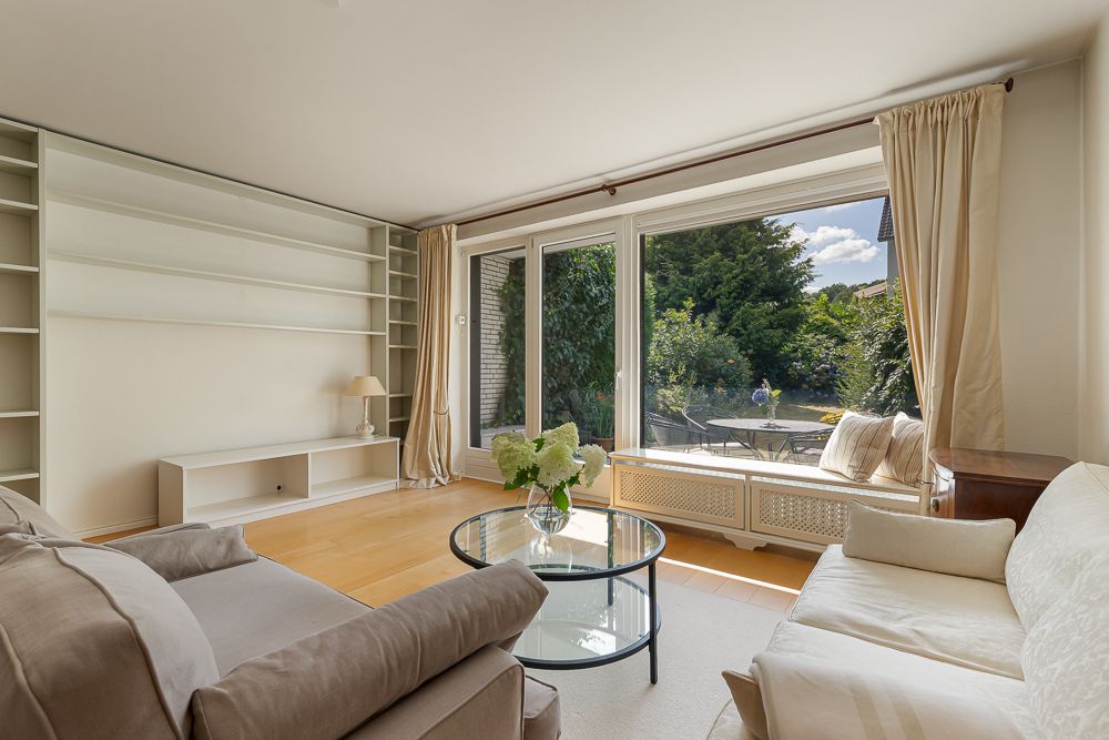 Helles Wohnzimmer mit zwei Sofas, Glastisch, Regalen und großen Fenstern zum Garten.