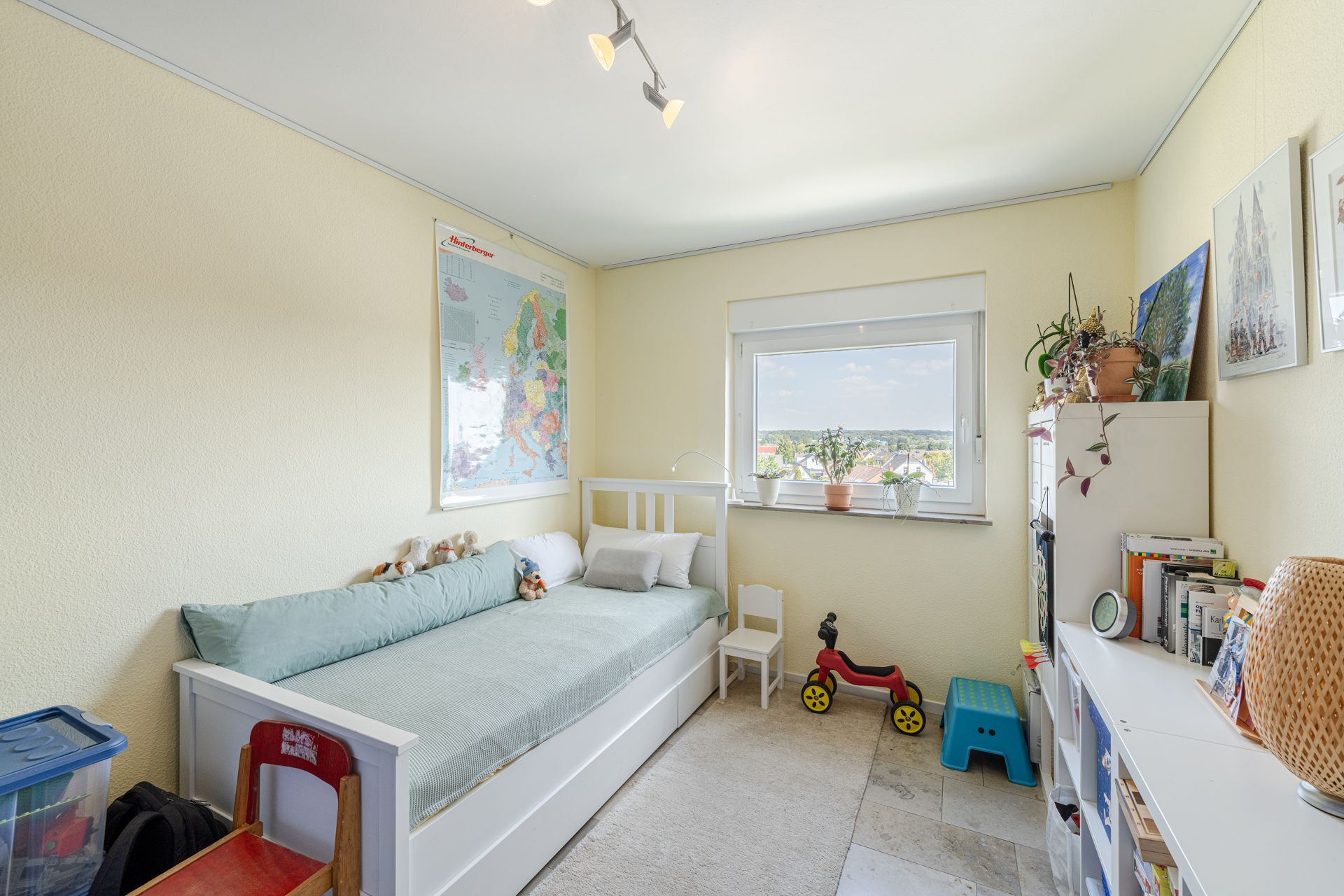 Helles Kinderzimmer mit Einzelbett, Spielzeug, Bücherregal und großem Fenster.