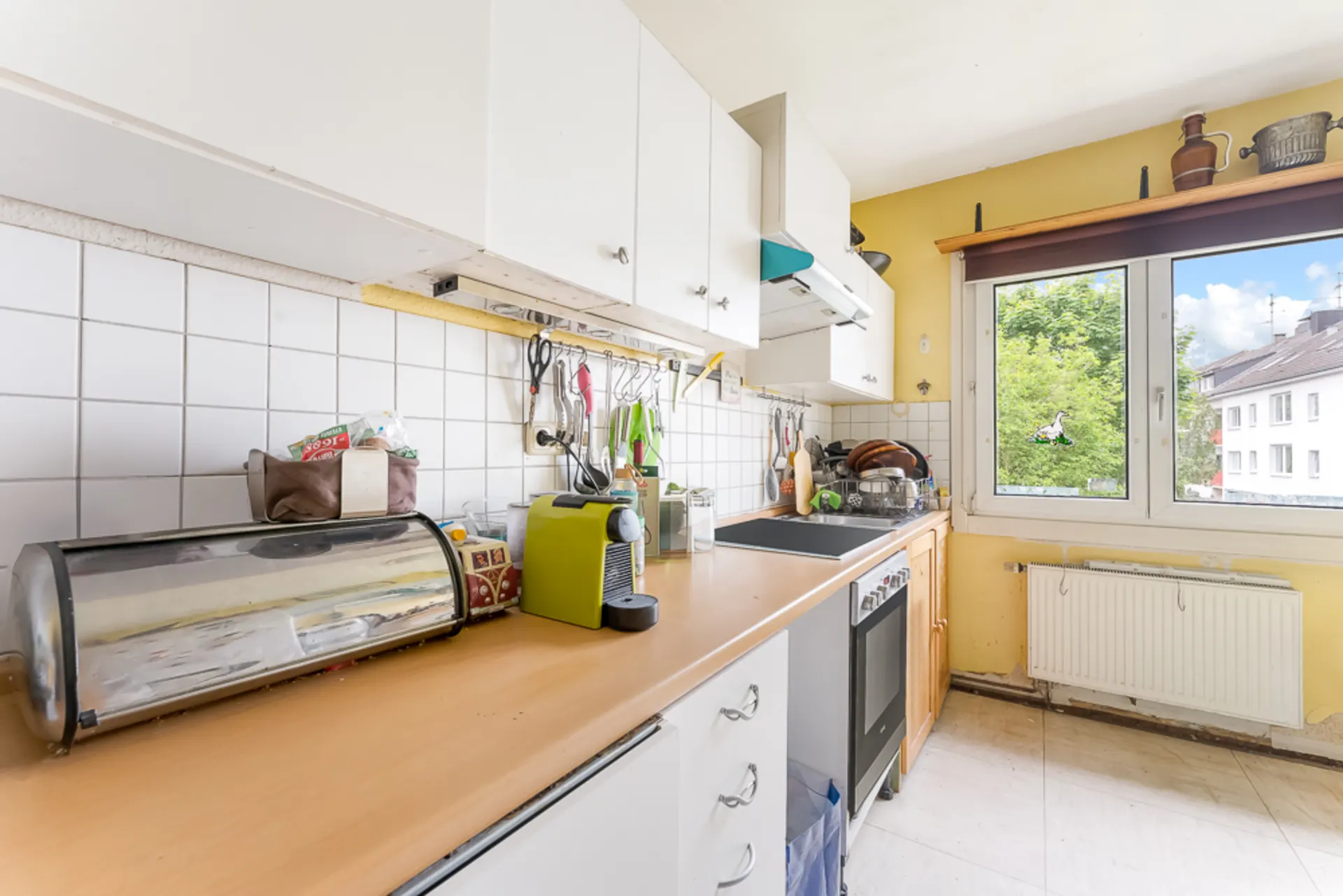 Helle Küche mit weißen Schränken, Kaffeemaschine, Toaster und Fenster mit Blick nach draußen.