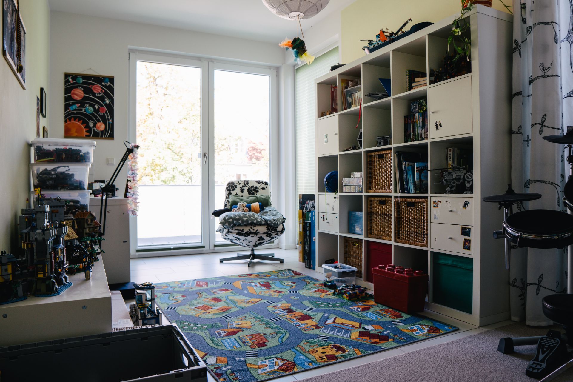 Helles Kinderzimmer mit Regalwand, Spielteppich und großem Fenster mit Blick nach draußen.