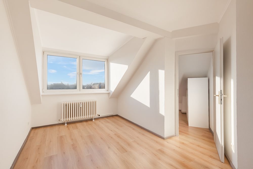 Helles Zimmer mit Dachschräge, Fenster, Heizkörper und Holzfußboden.