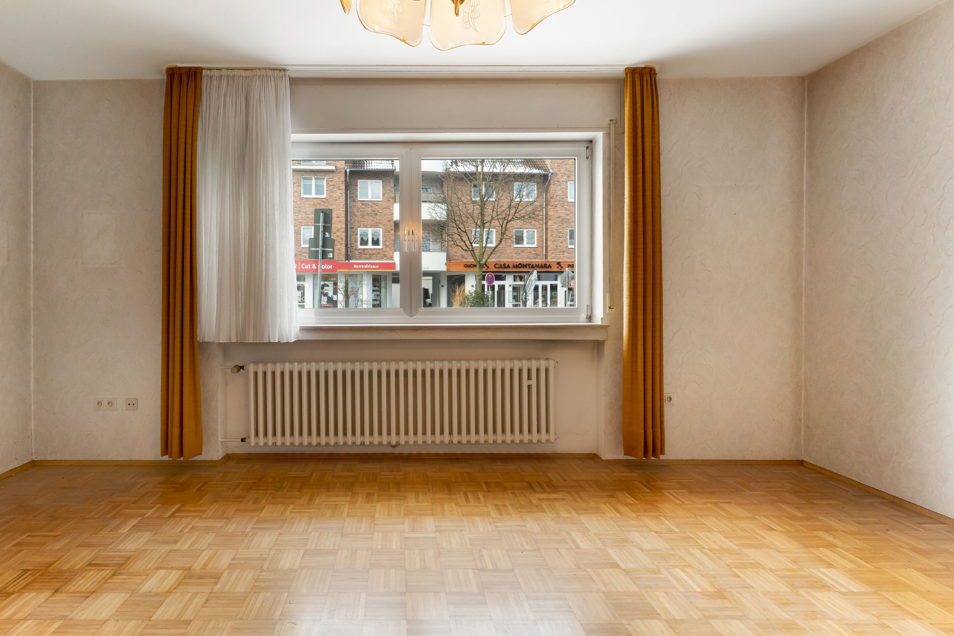 Leeres Zimmer mit Parkettboden, großem Fenster, Heizkörper und gelben Vorhängen.