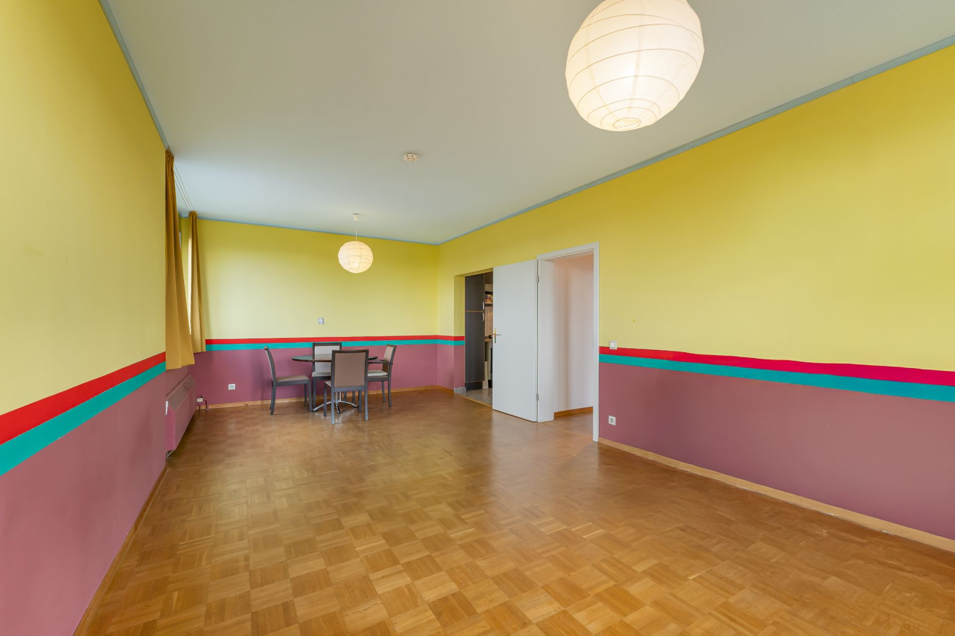 Großes Zimmer mit bunten Wänden, Holzboden, Esstisch und Stühlen, mit zwei runden Deckenlampen.