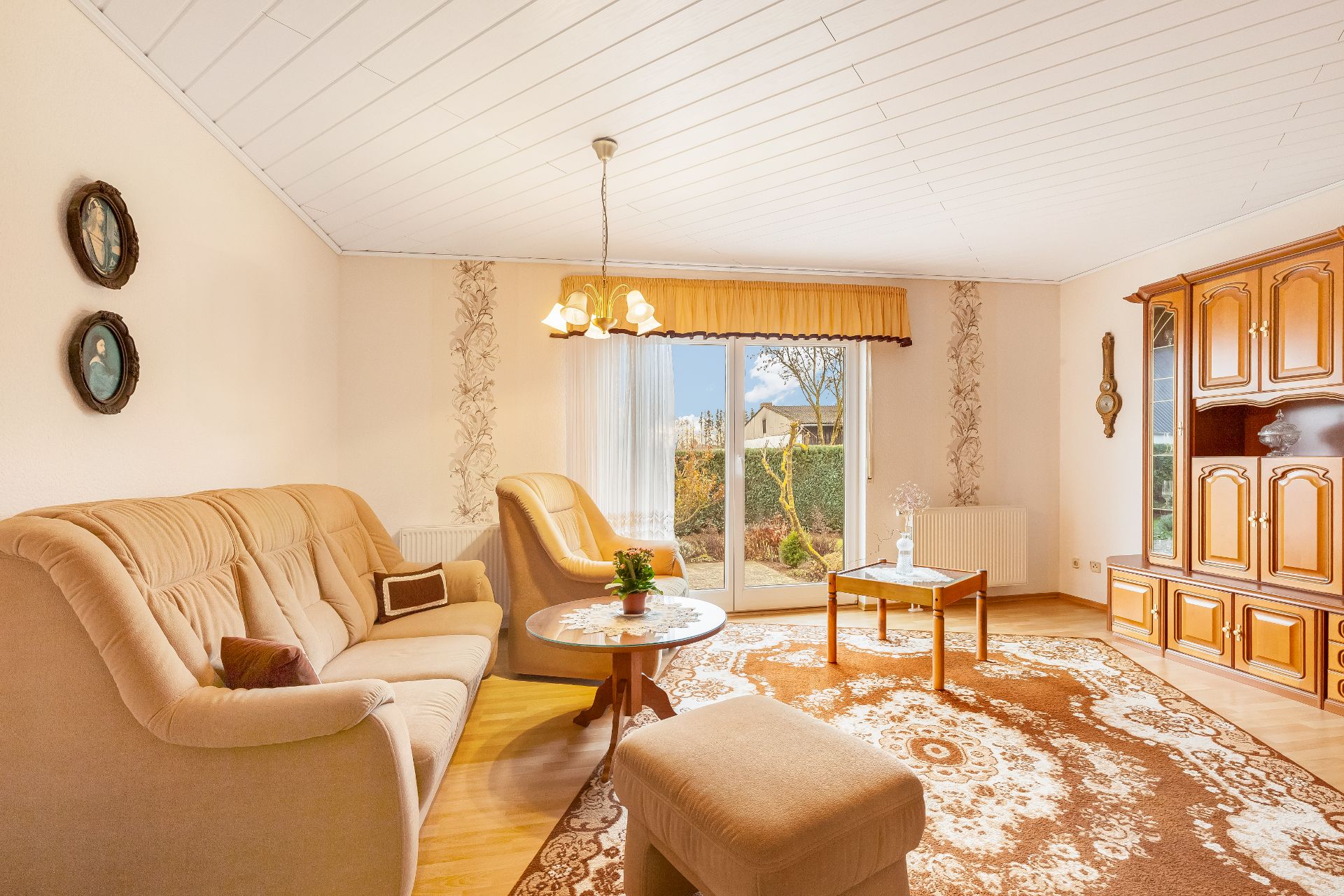 Helles Wohnzimmer mit beigen Polstermöbeln, Teppich, Holzmöbeln und großem Fenster zum Garten.