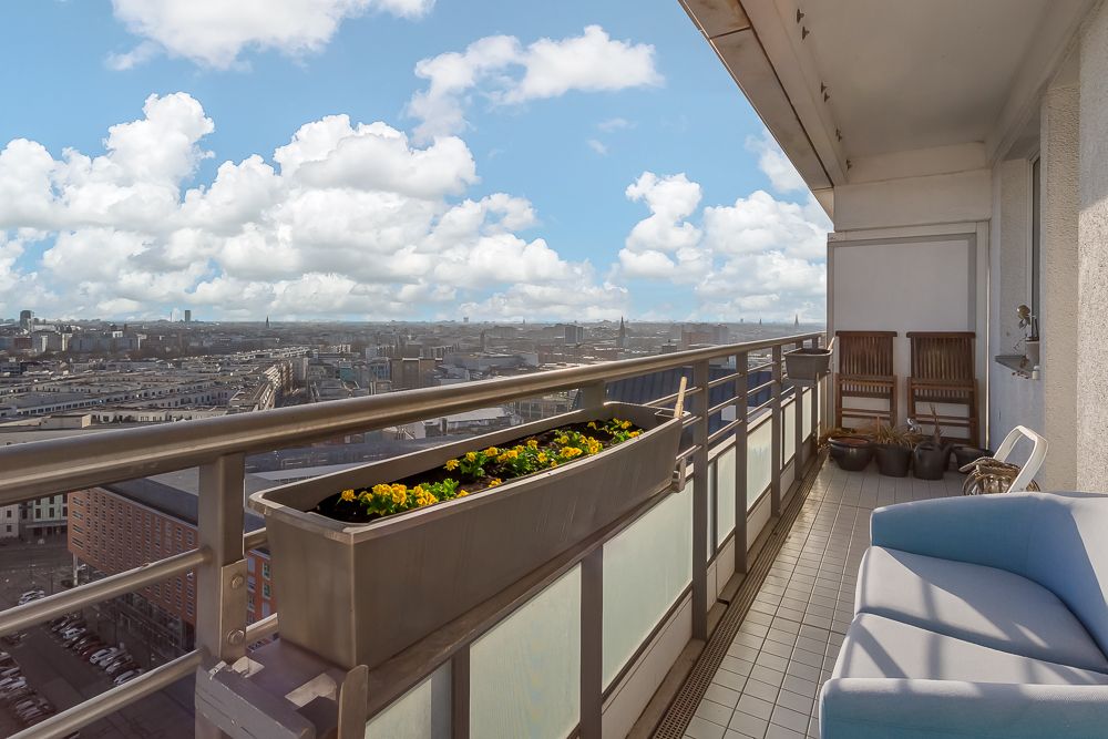 Balkon mit Blumenkasten, Blick über die Stadt, zwei Stühle, Blumentöpfe und ein Sofa.