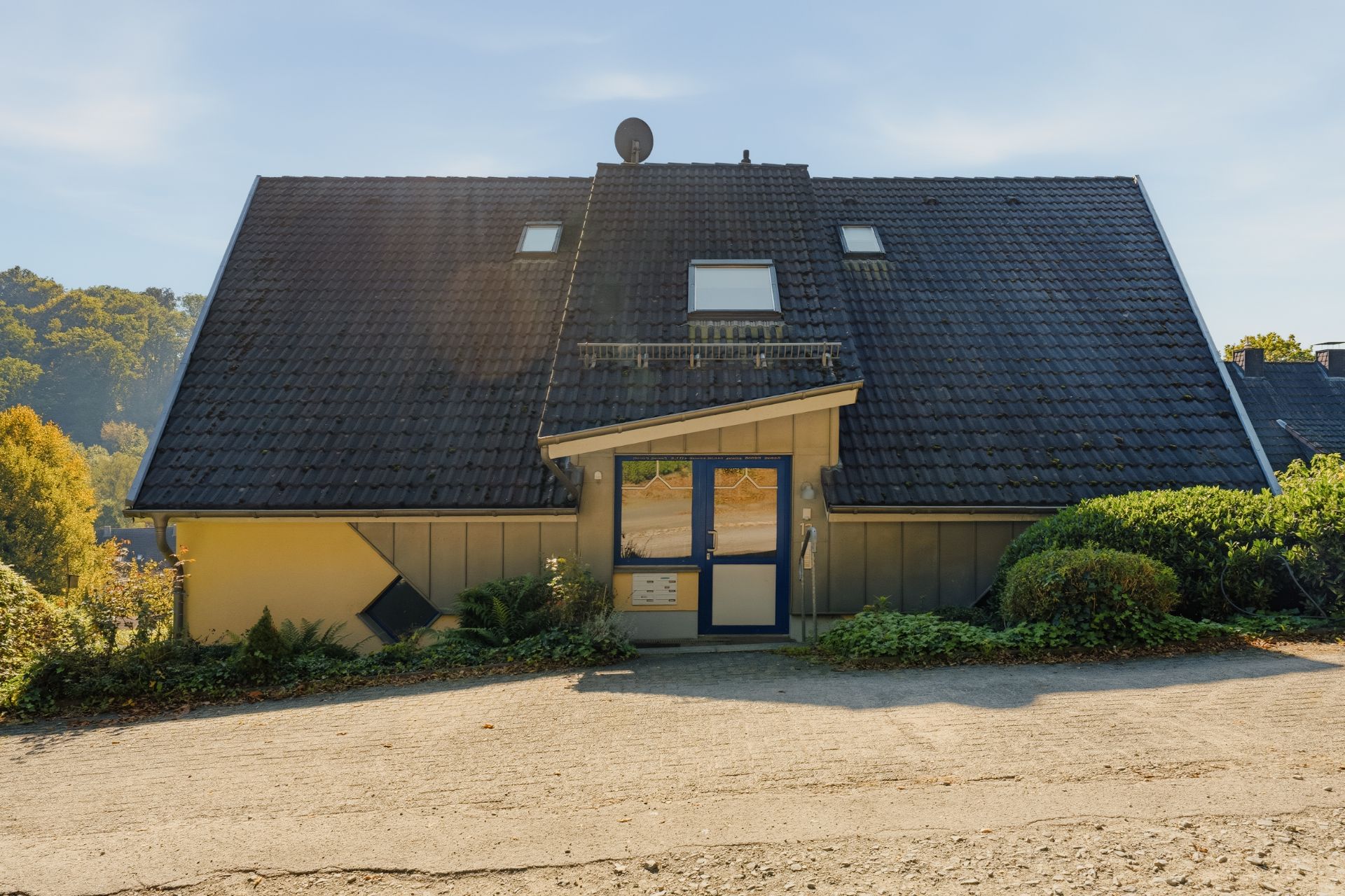 Haus mit dunklem Satteldach, Eingangsbereich und umgebenden Büschen, vorne Zufahrtsweg.