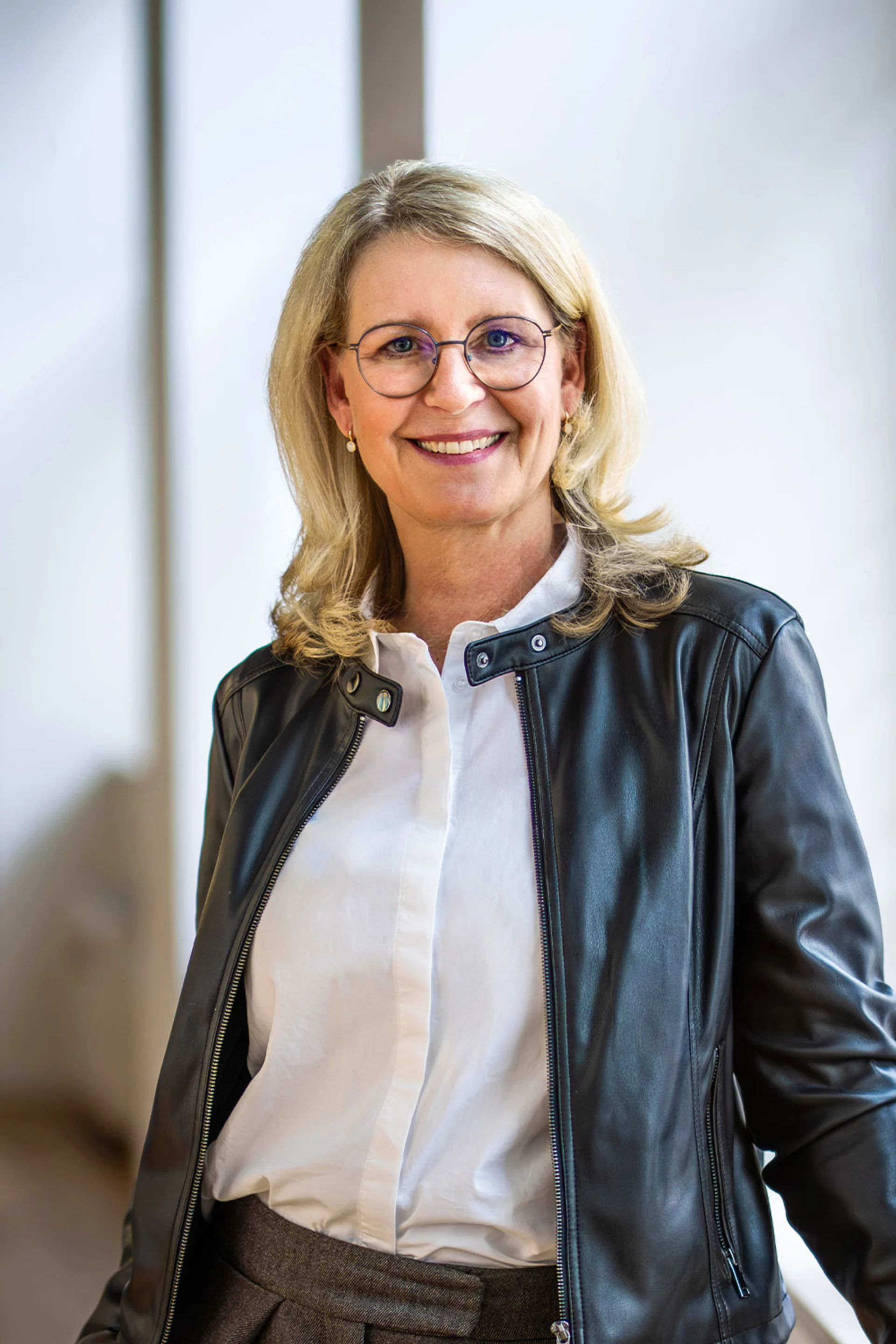 Frau mit blonden Haaren, Brille und schwarzer Lederjacke steht in einem hellen Raum.