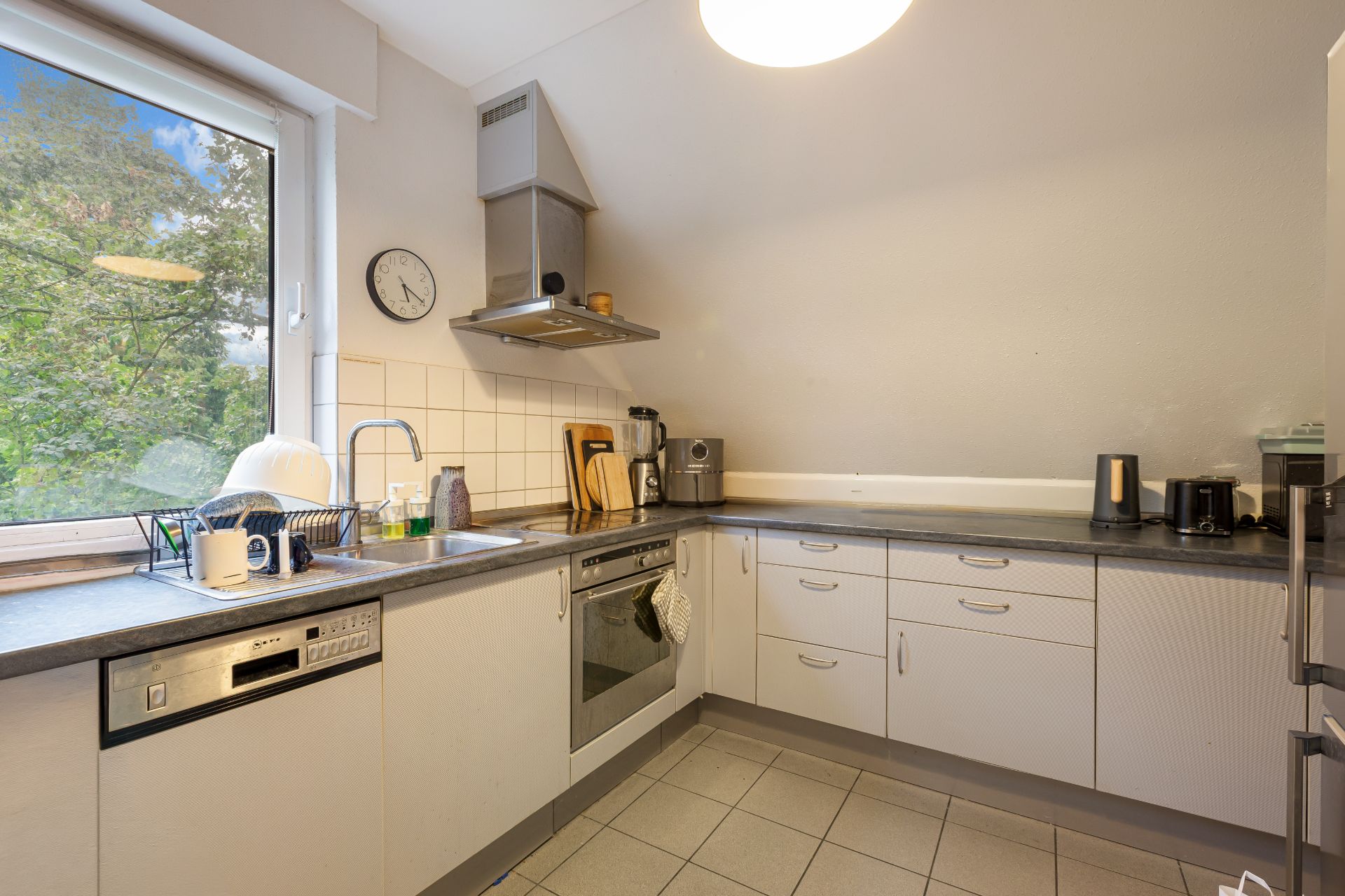 Moderne Einbauküche mit weißen Schränken, großem Fenster, Spüle und diversen Küchengeräten.