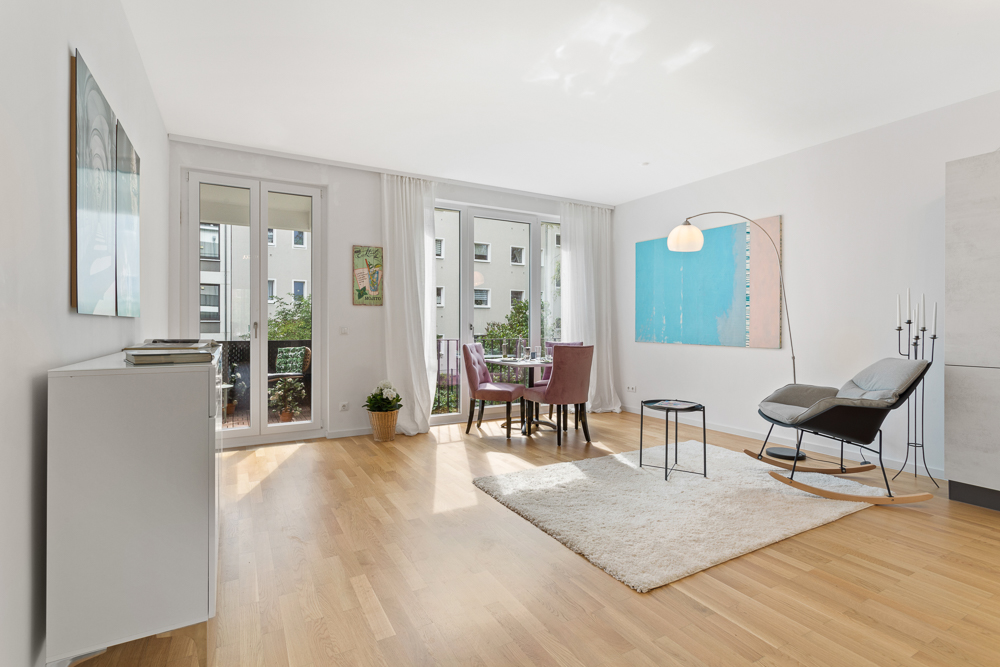 Helles Wohnzimmer mit Holzboden, Esstisch, Sessel, Teppich und Zugang zum Balkon.