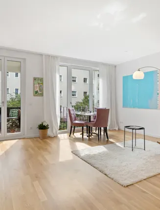 Helles Wohnzimmer mit Holzboden, Esstisch, Sessel, Teppich und Zugang zum Balkon.