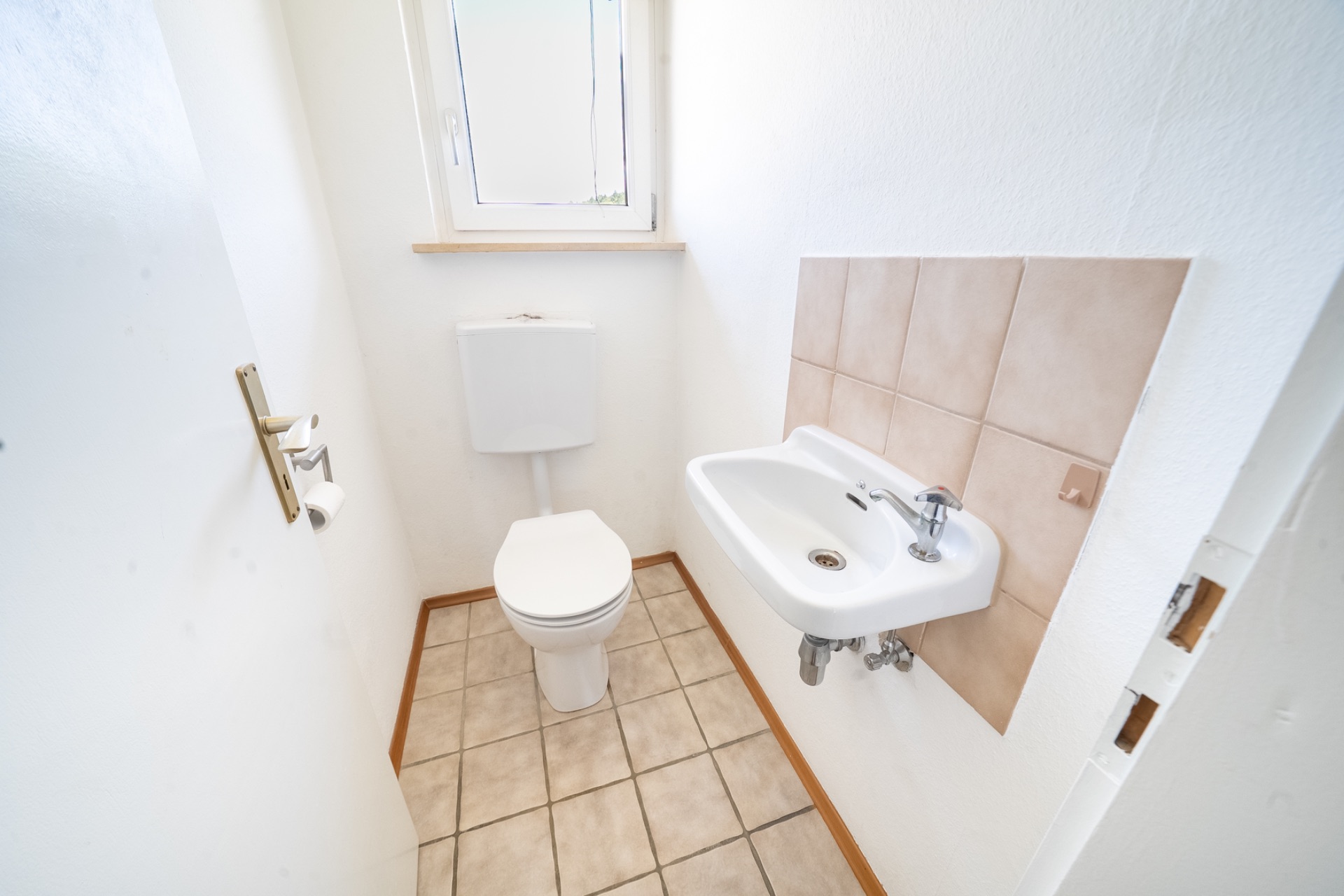 Kleines Badezimmer mit WC, Waschbecken und Fenster, weiße Wände und gefliester Boden.