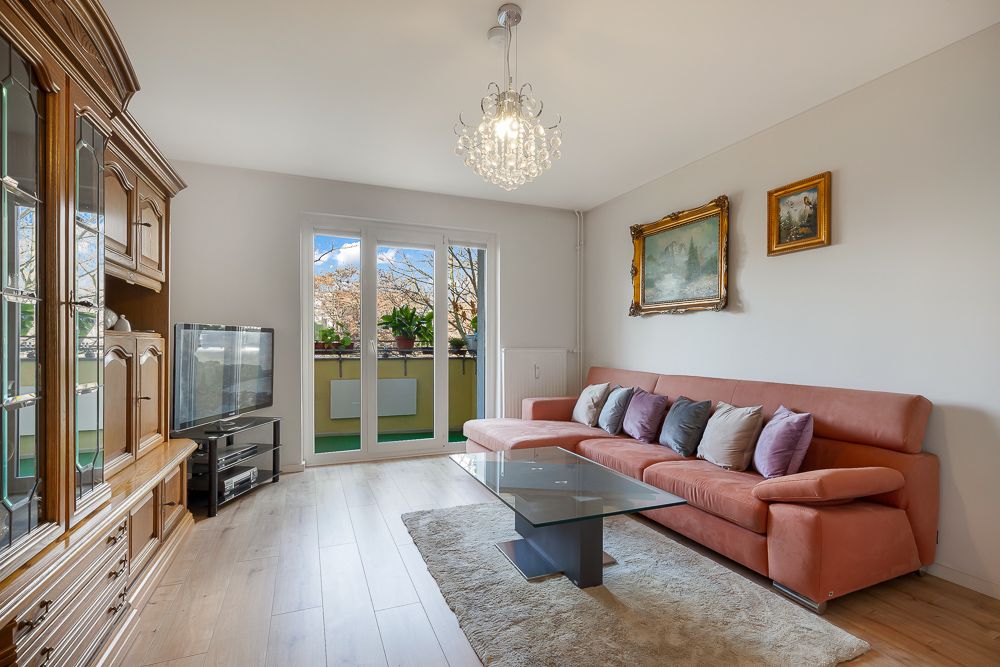 Helles Wohnzimmer mit Ecksofa, Glastisch, Teppich, TV und Zugang zu einem Balkon.