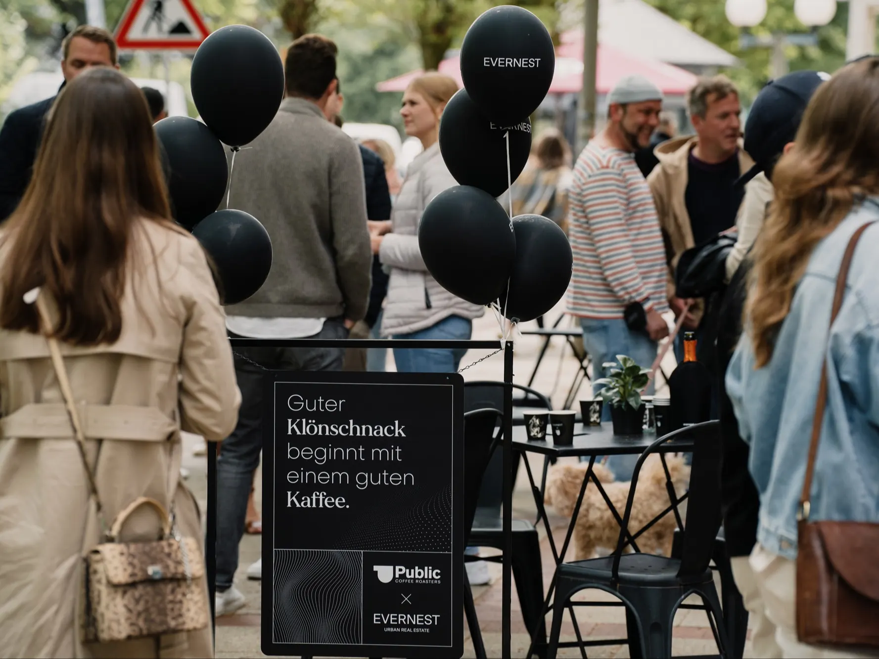 Menschen stehen und sitzen an Tischen im Freien, schwarze Ballons und ein Schild mit Text sind sichtbar.