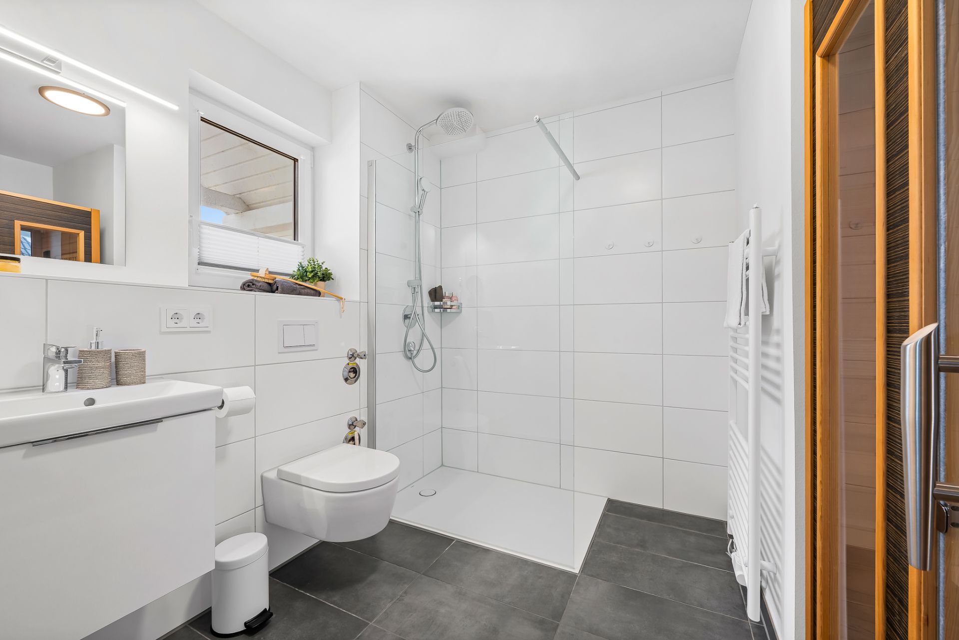 Modernes Badezimmer mit ebenerdiger Dusche, Waschbecken und WC, weißen Fliesen und grauem Boden.