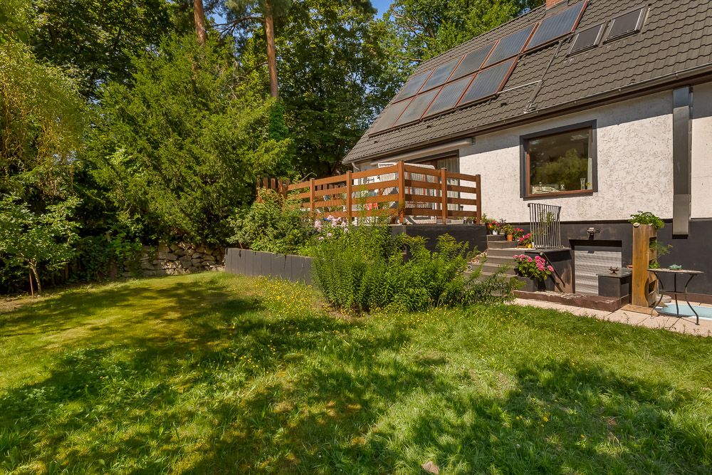 Garten mit Rasenfläche, Terrasse mit Holzgeländer und Haus mit Solarpanelen auf dem Dach.