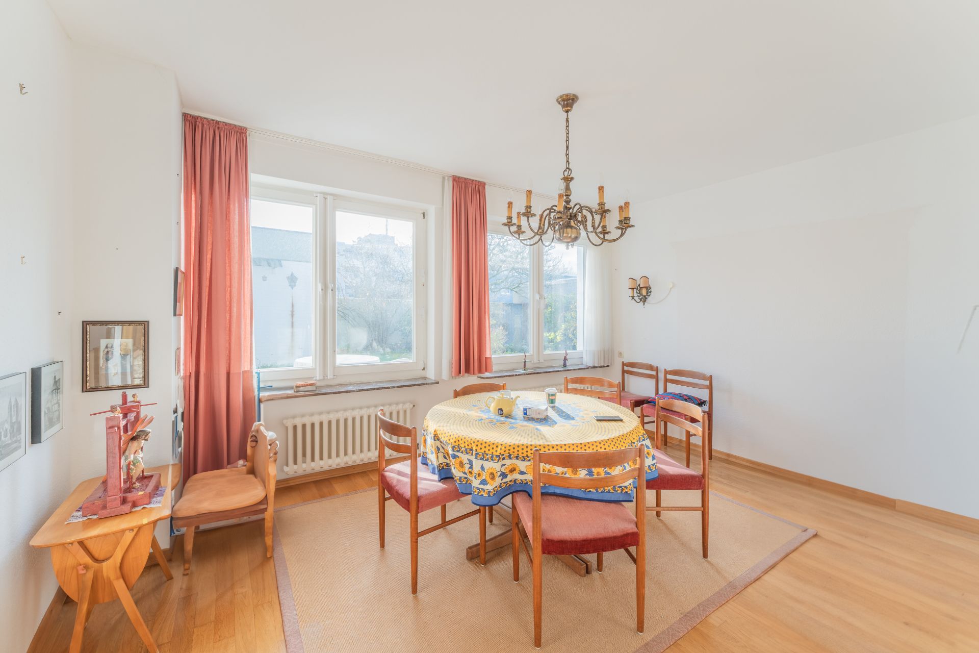 Helles Esszimmer mit rundem Tisch, sechs Stühlen, Kronleuchter und großen Fenstern mit roten Vorhängen.
