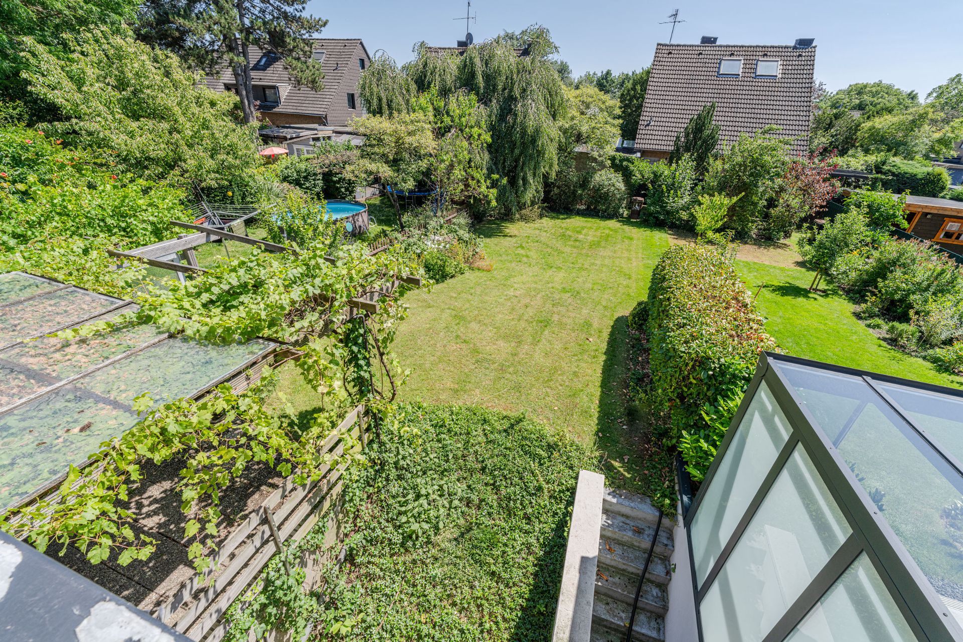 Großer Garten mit Rasenfläche, Bäumen, Sträuchern und überdachter Terrasse, Blick von oben.