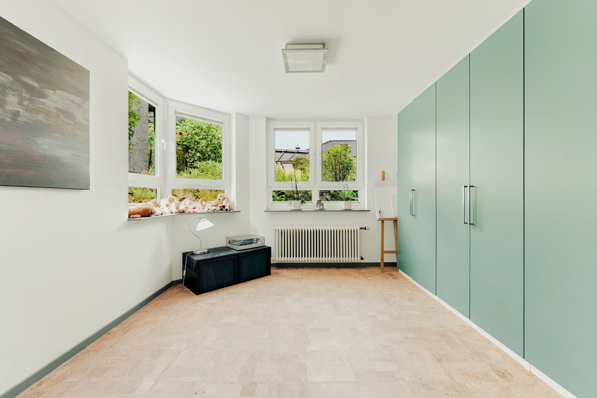 Helles Zimmer mit großen Fenstern, grünen Einbauschränken und Deko auf der Fensterbank.