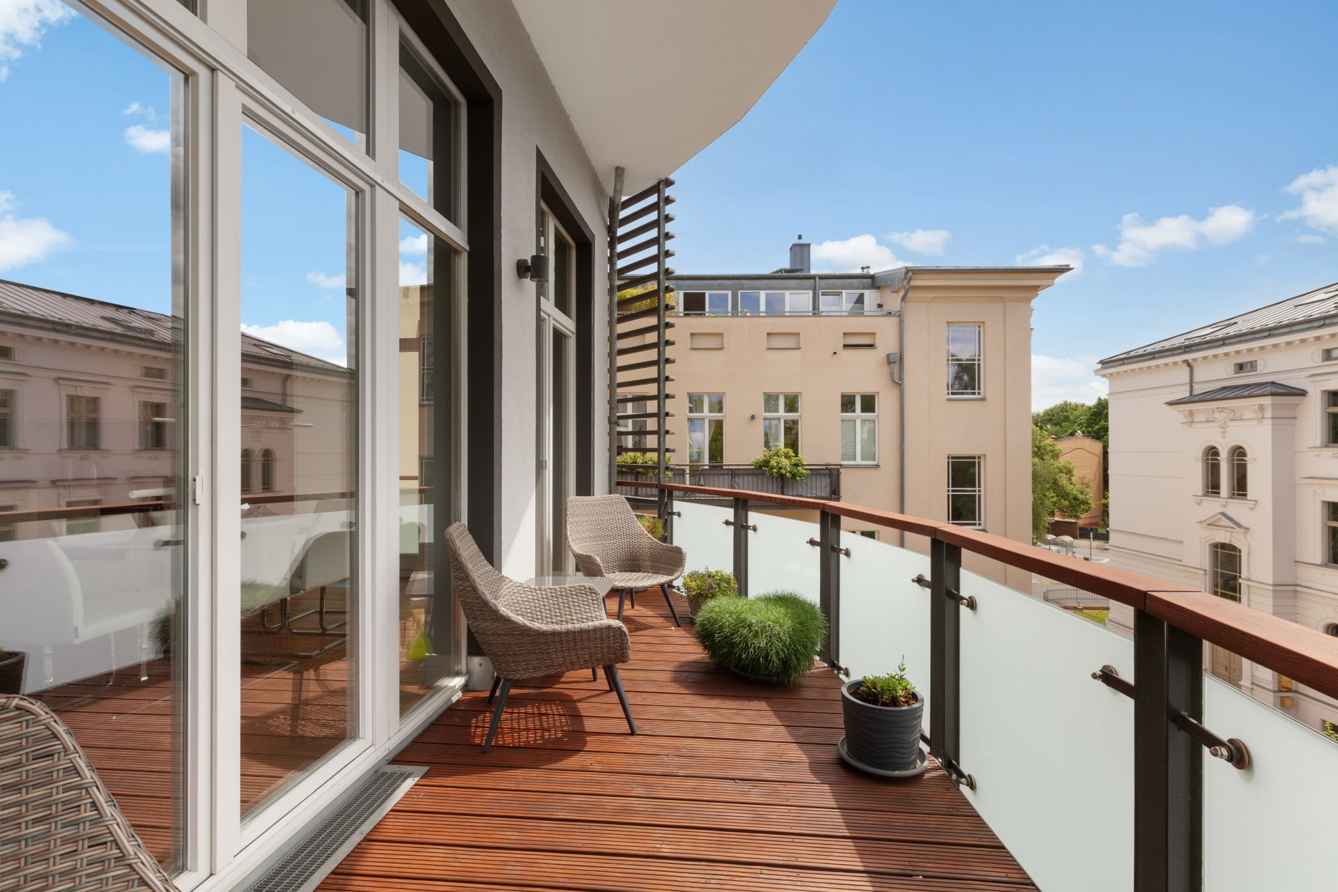 Balkon mit Holzboden, zwei Stühlen, Pflanzen und Blick auf benachbarte Gebäude.