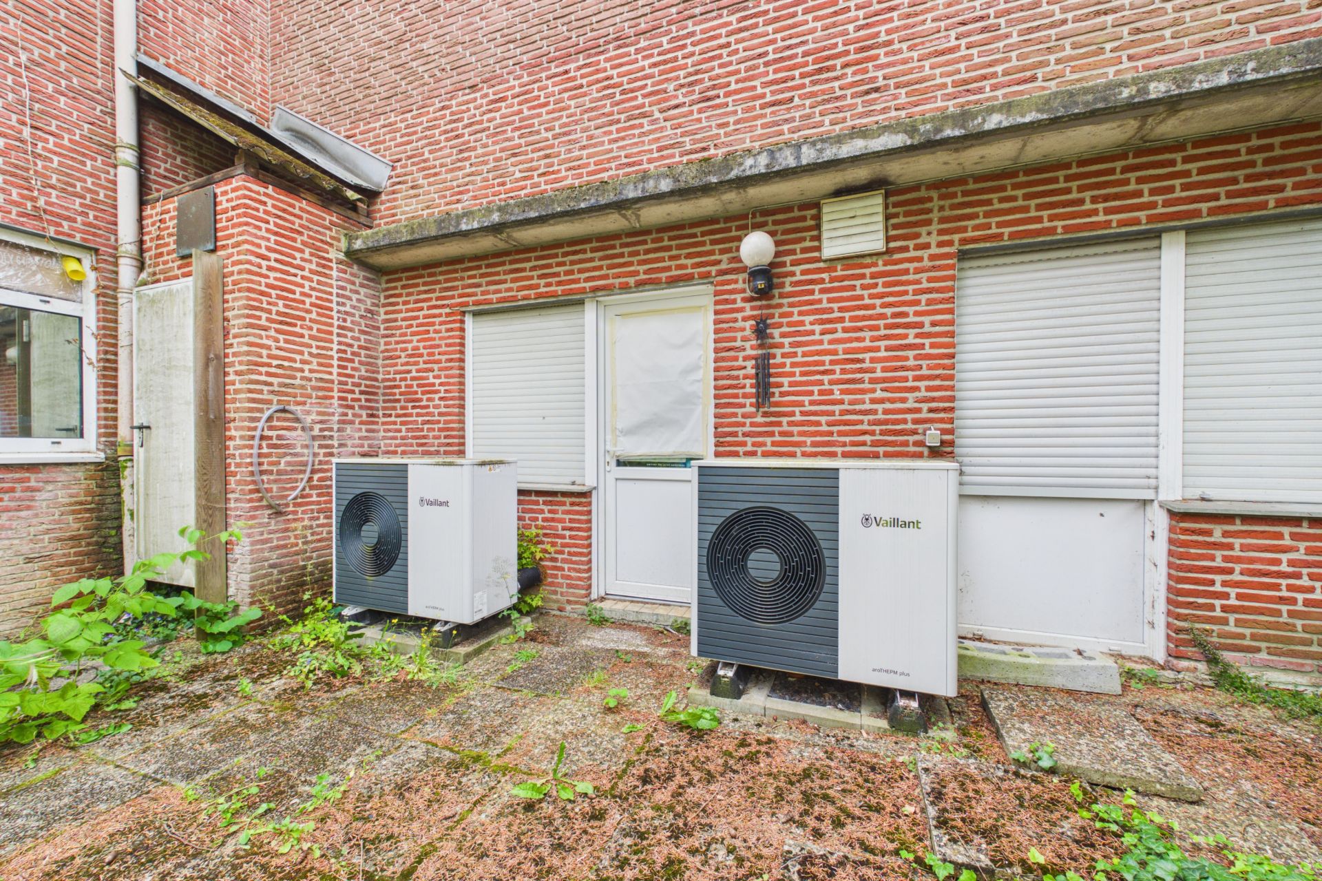 Zwei Wärmepumpen stehen auf einer Terrasse vor einer rot geklinkerten Hauswand mit weißen Fenstern und Türen.