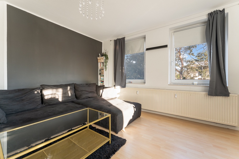 Wohnzimmer mit dunklem Sofa, Glastisch, zwei Fenstern und hellem Holzboden.