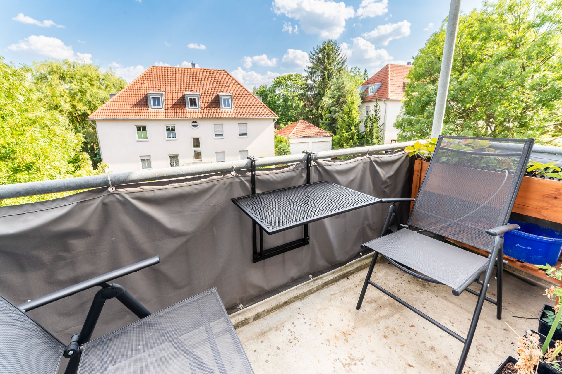 Kleiner Balkon mit zwei Stühlen, kleinem Tisch und Blick auf Wohnhäuser und Bäume.