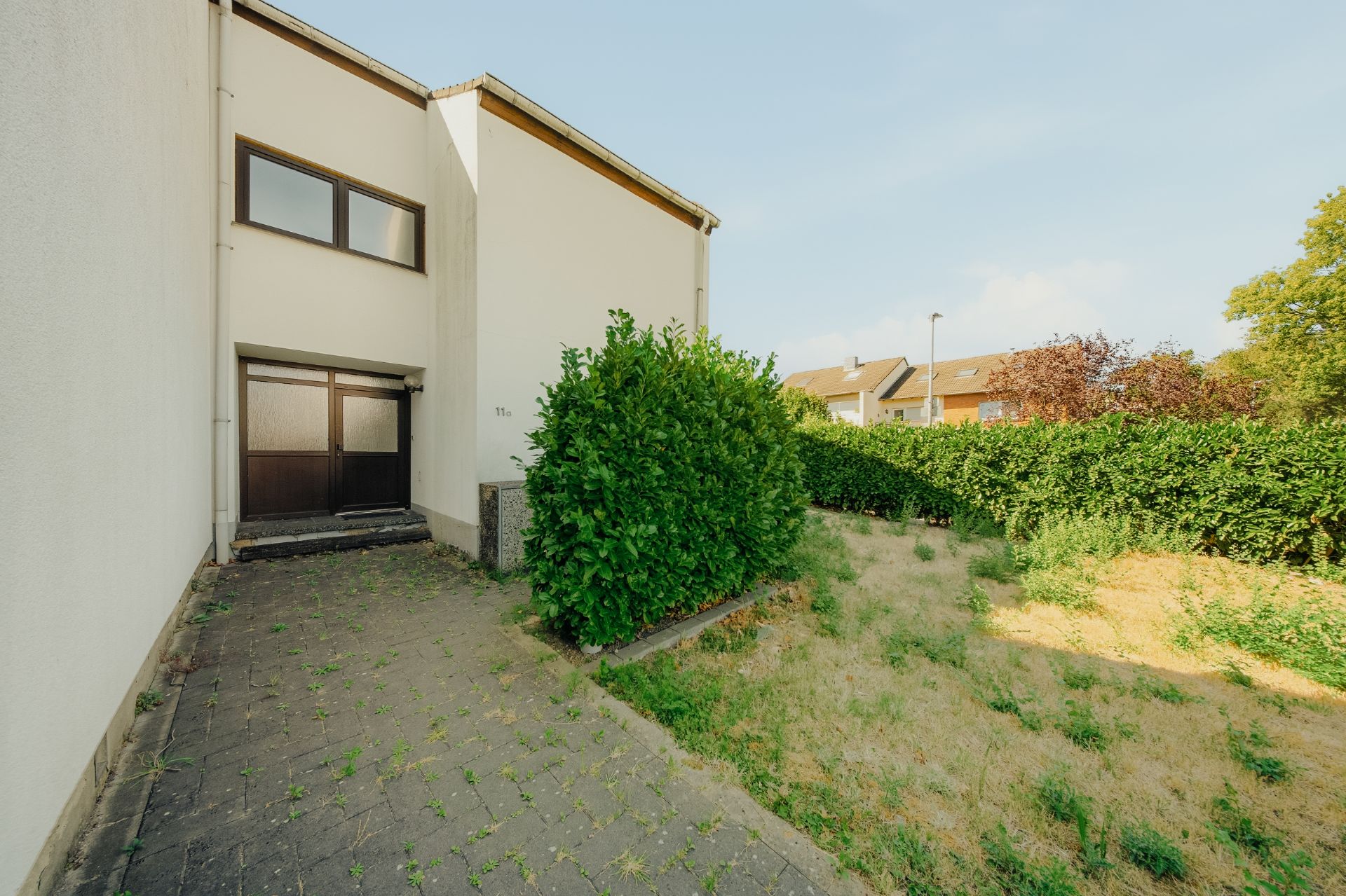 Eingangsbereich eines Wohnhauses mit Pflasterweg und Rasenfläche, umgeben von Hecken.