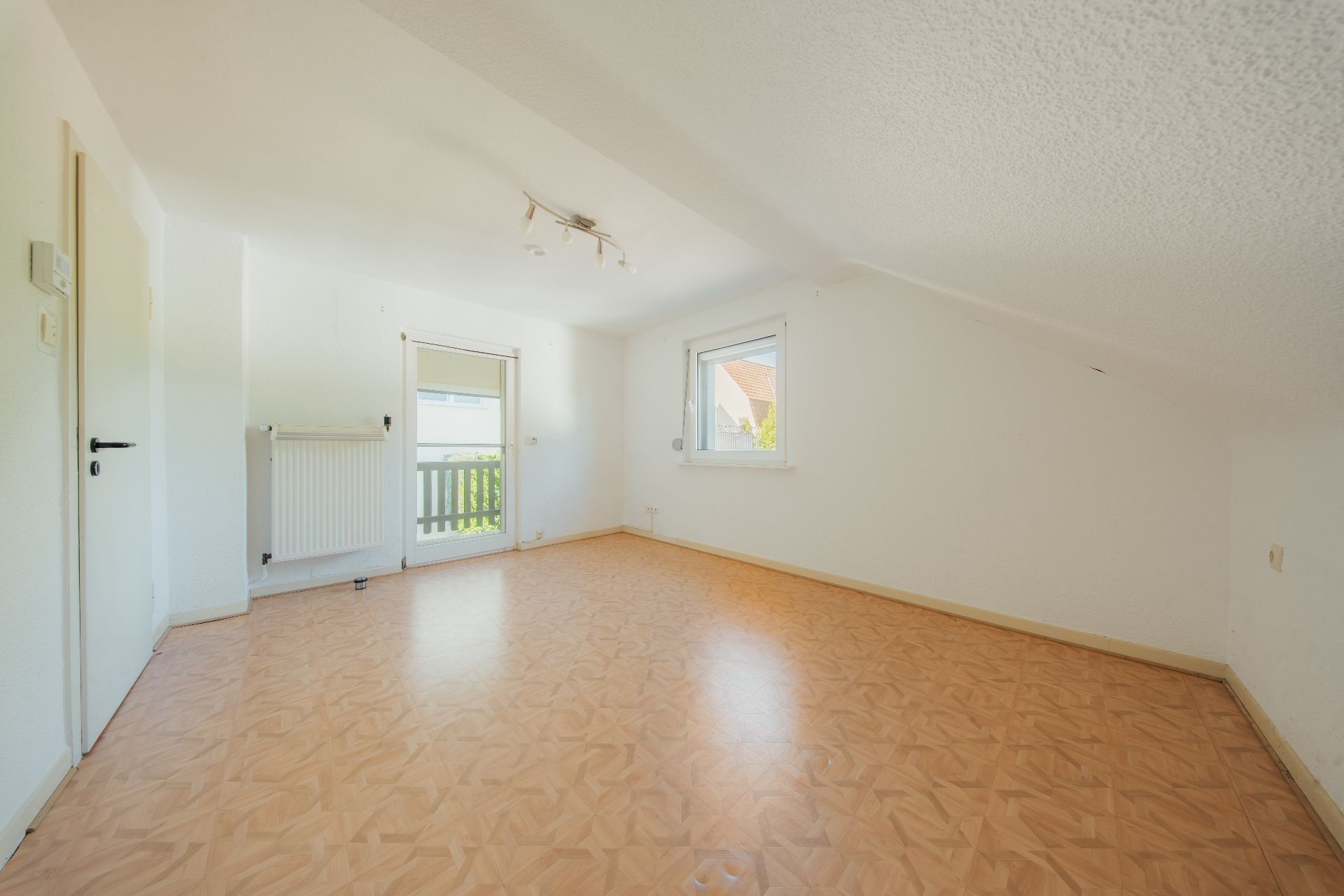 Helles, leeres Zimmer mit Dachschräge, Holzboden, Fenster und Tür zu einem kleinen Balkon.