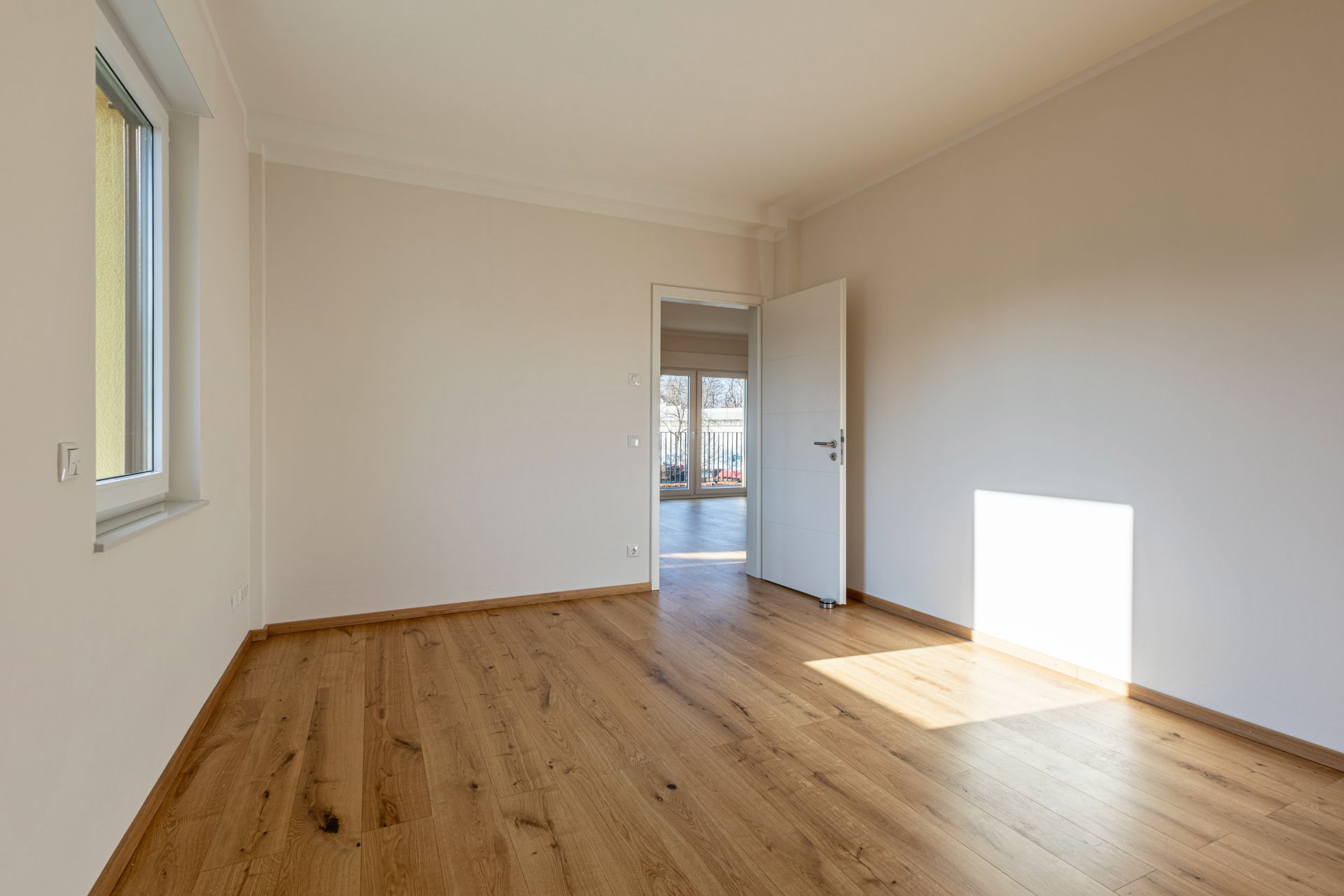 Leeres Zimmer mit Holzboden, weißer Wand, Fenster und geöffneter Tür.