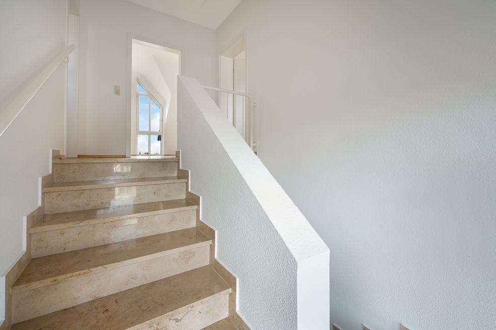 Treppe mit beigen Marmorstufen und weißem Geländer in einem hellen Flur.