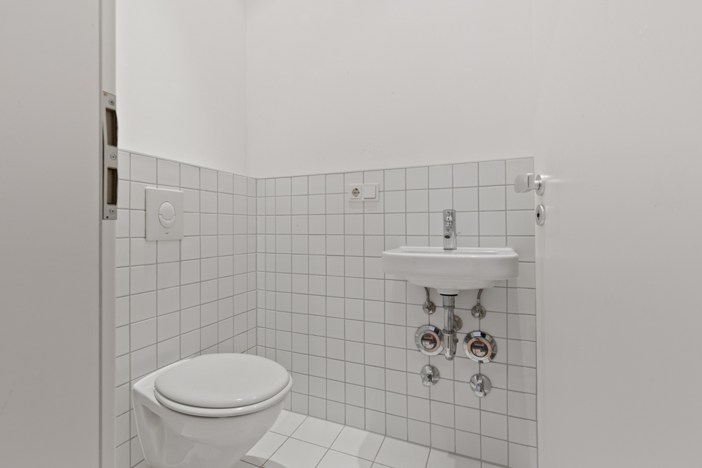 Kleines Badezimmer mit weißem WC und kleinem Waschbecken, weiße Fliesen an Wänden und Boden.
