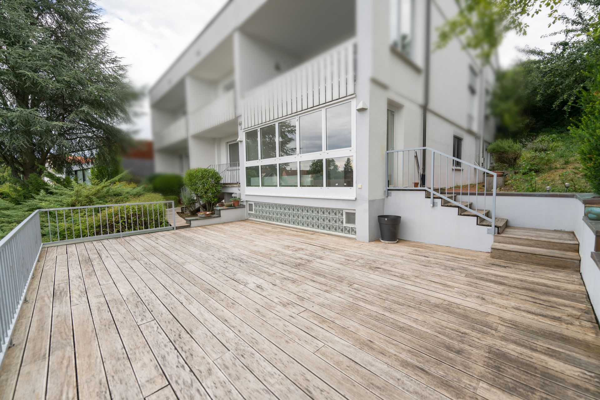 Große Holzterrasse mit Geländer vor einem modernen Wohnhaus, umgeben von Grünpflanzen.