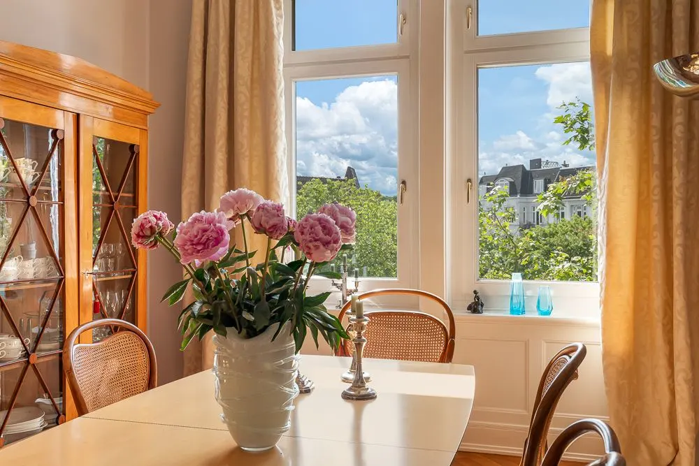Esszimmer mit Holztisch, Blumen in Vase, Vitrine und Blick aus dem Fenster auf Bäume und Gebäude.
