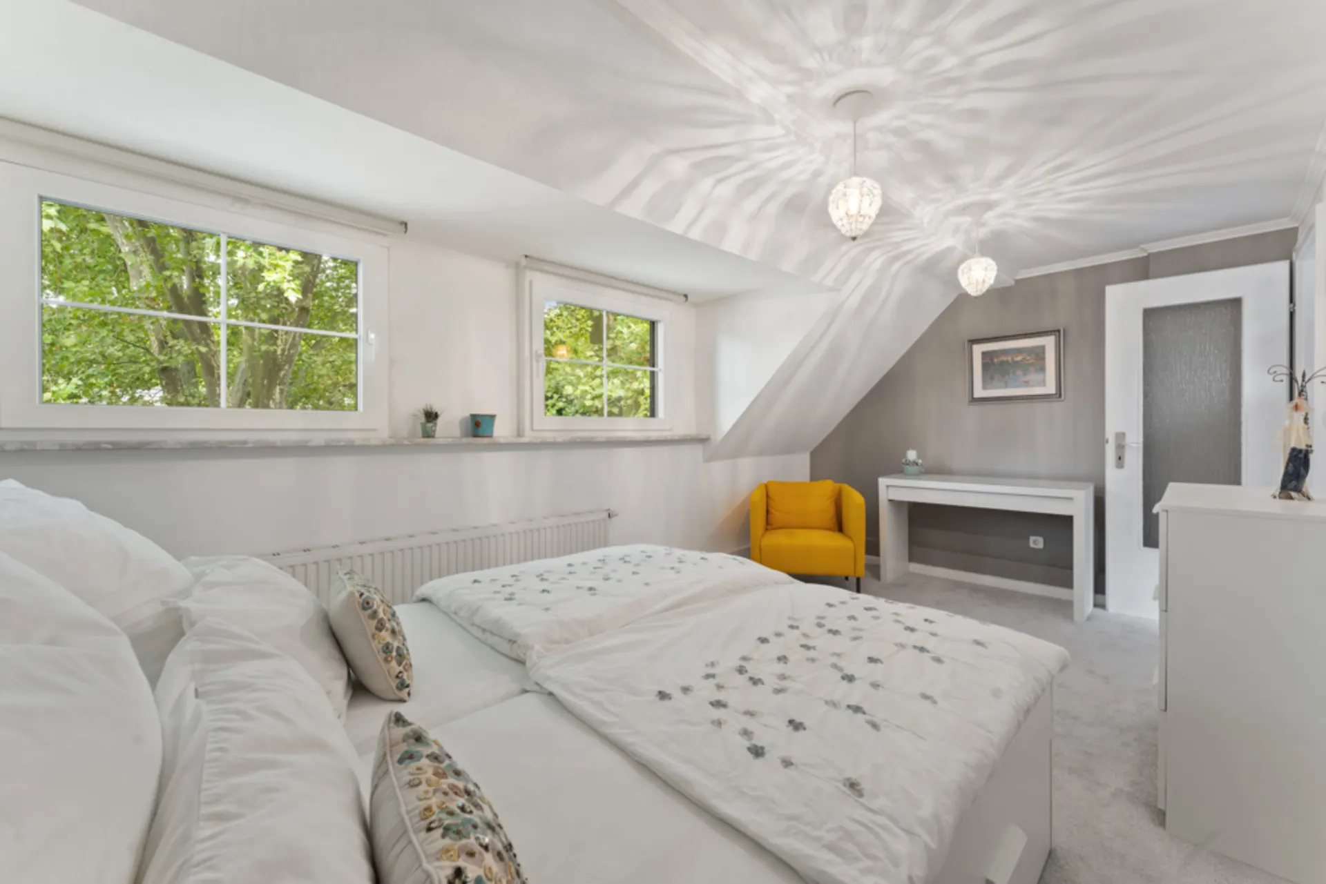 Helles Schlafzimmer mit Doppelbett, gelbem Sessel und zwei Fenstern mit Blick auf Bäume.