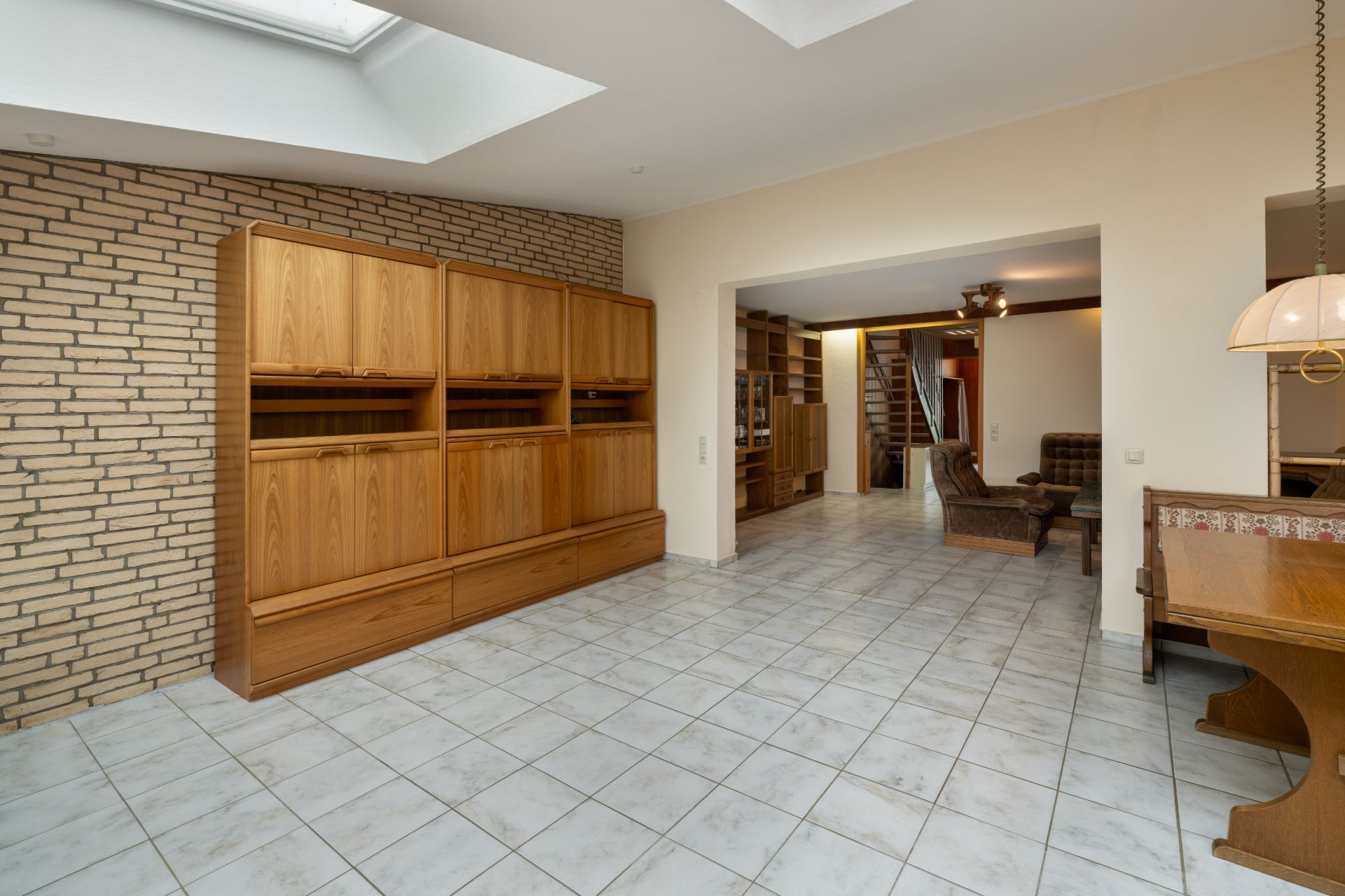 Heller Raum mit Fliesenboden, Holzschrank, Esstisch und offener Durchgang zum Wohnzimmer.